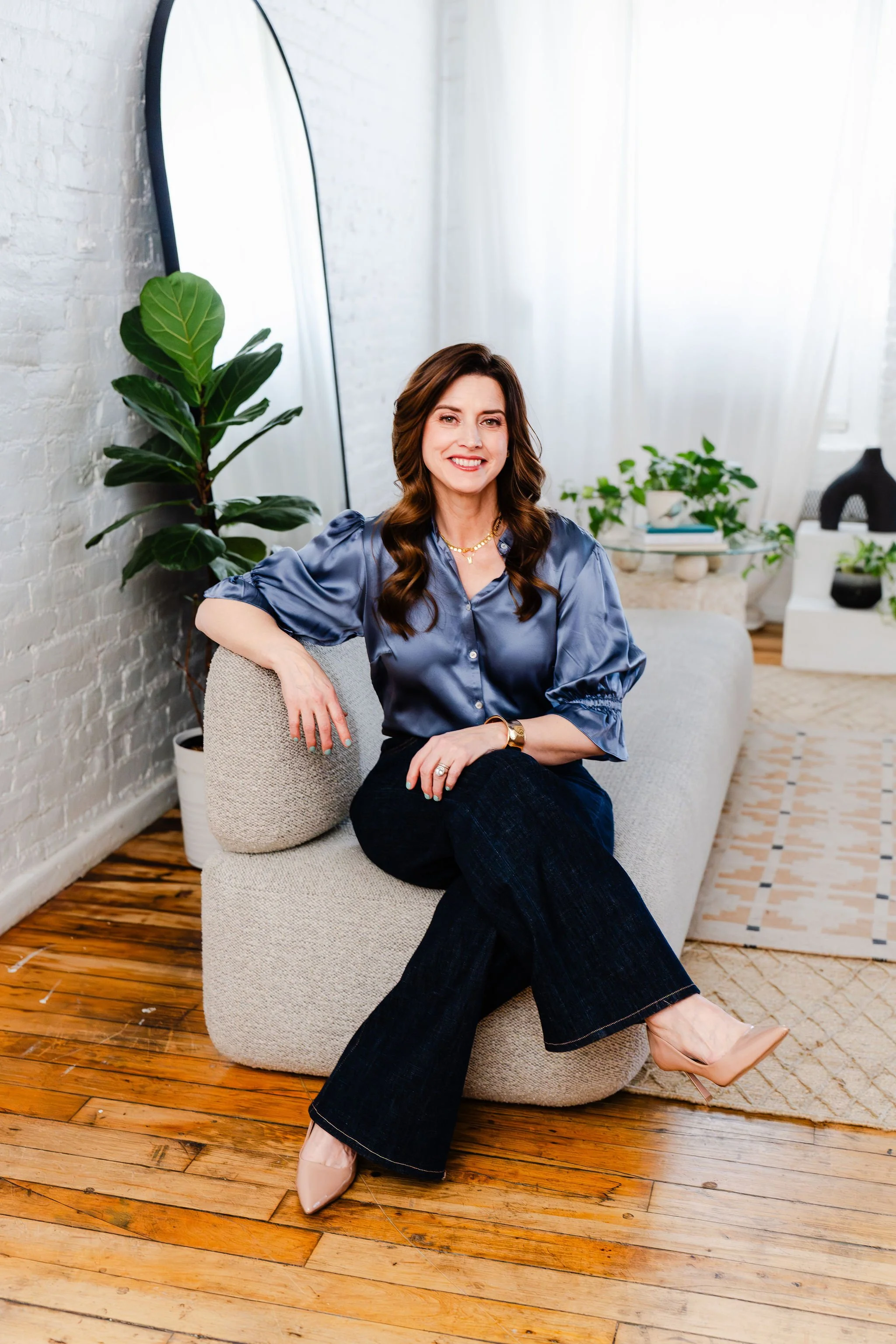 A woman with long dark hair, wearing a satin blue blouse, black jeans, and nude high heels, sitting on a beige sofa in a bright room with white brick walls, wooden floors, and large windows with white curtains. There are green plants and decorative items in the background.