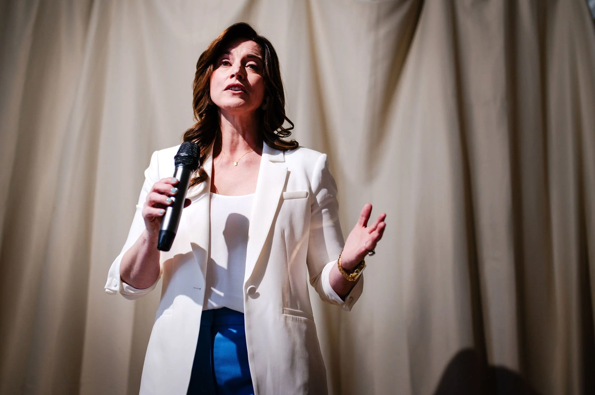 A woman with dark brown hair, wearing a white blazer and matching top, holding a microphone, standing in front of a beige curtain, speaking or presenting.