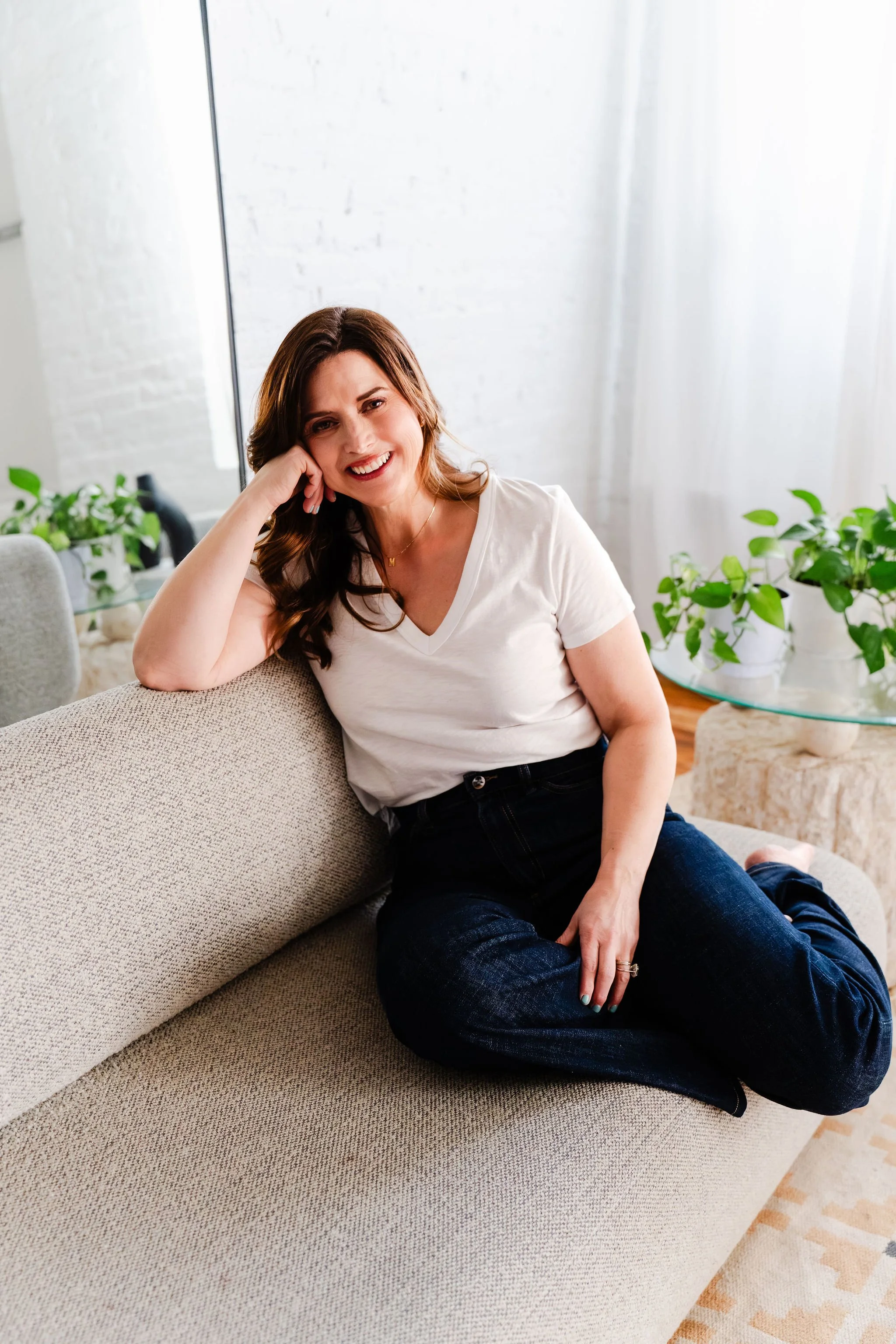 A woman with brown hair, wearing a white t-shirt and dark jeans, sitting on a beige sofa indoors, smiling and resting her head on her hand, with green plants and white brick wall in the background.