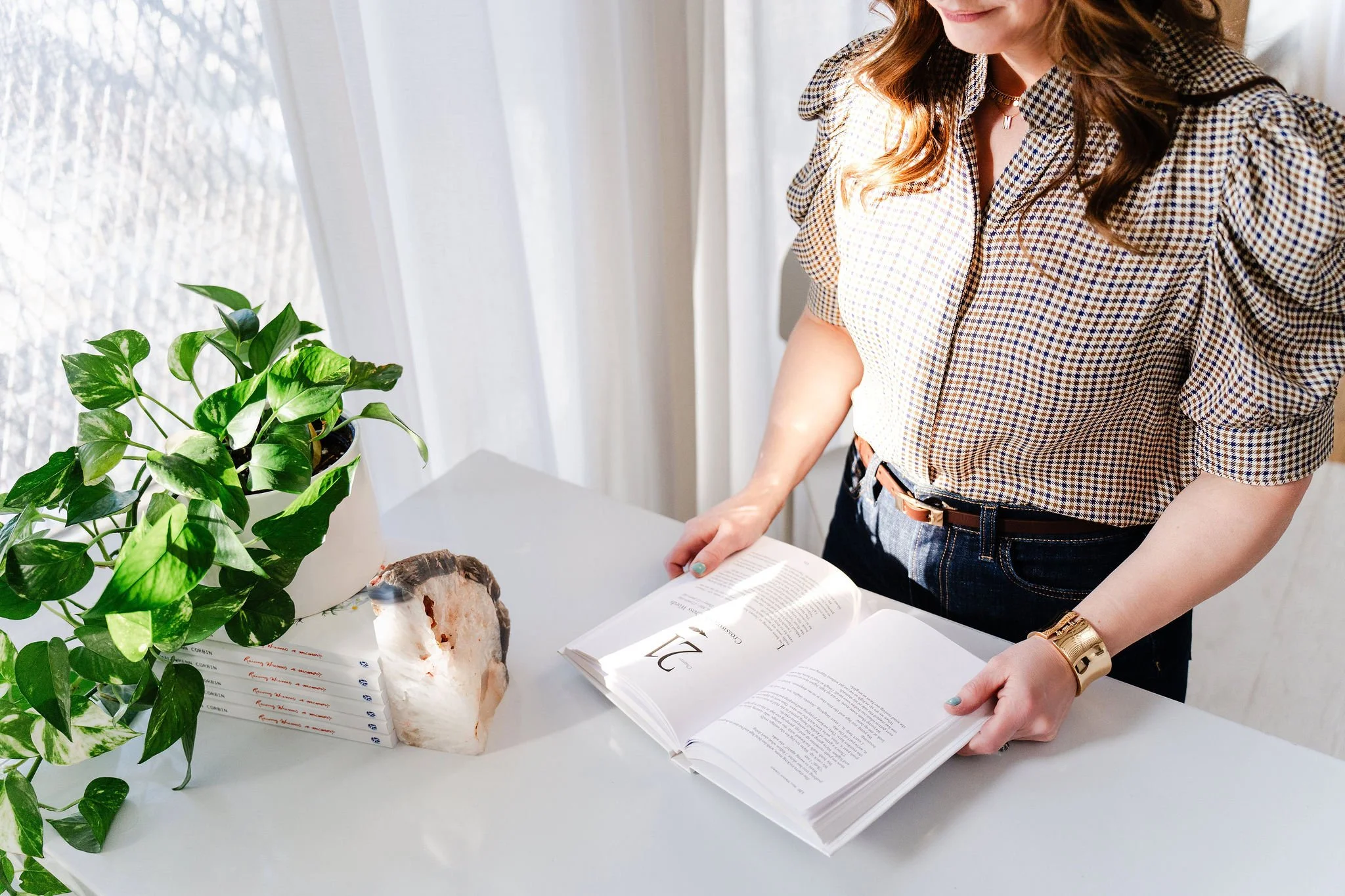 A woman with wavy brown hair wearing a checkered shirt, gold watch, and a letter necklace is standing at a white table with a green potted plant, a book, a large crystal, and a stack of small white papers with handwritten text.