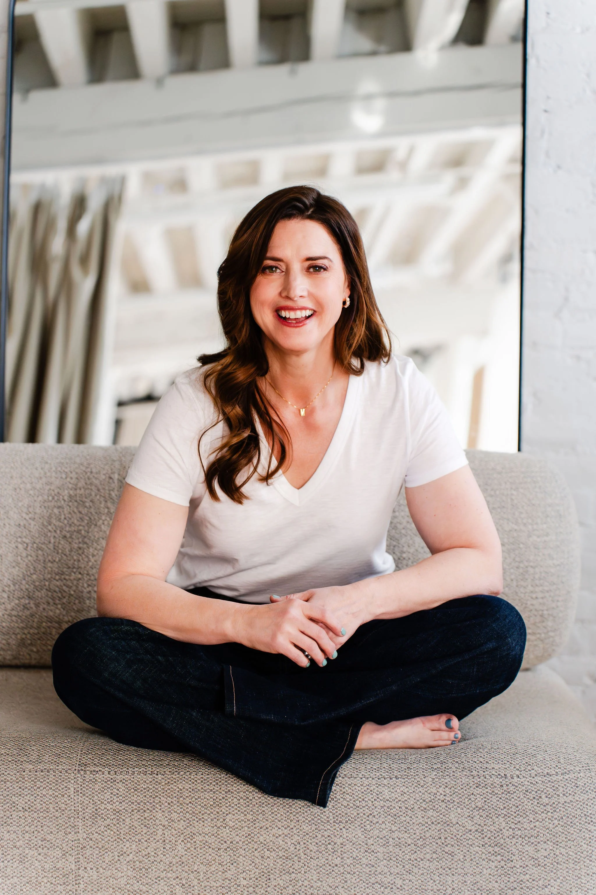 A woman with brown hair sitting cross-legged on a beige sofa with a textured fabric. She is smiling, wearing a white t-shirt, dark jeans, a gold necklace, and small earrings. The background features a large mirror reflecting white wooden beams and part of a white brick wall.