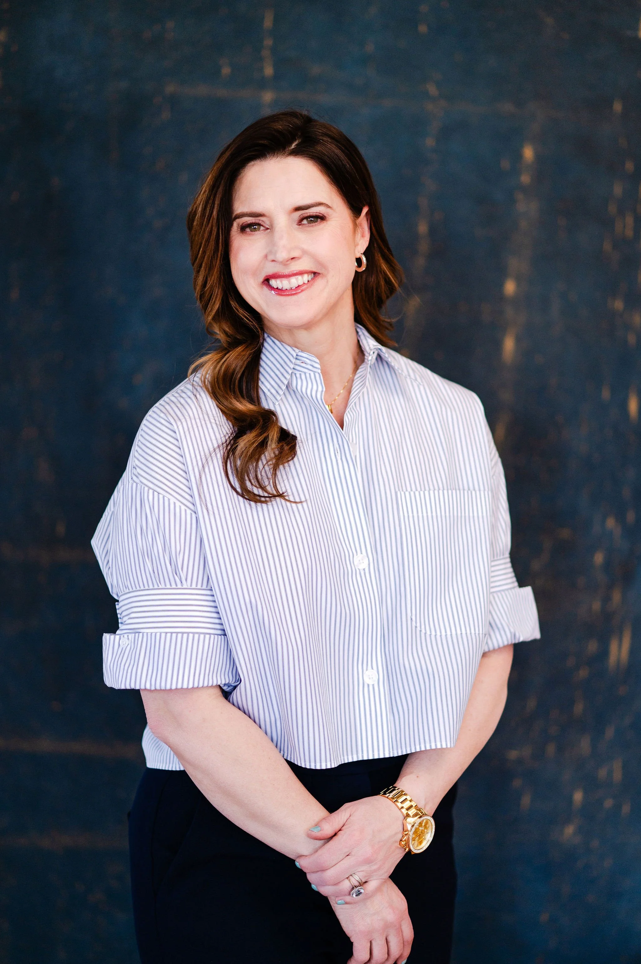 A woman with shoulder-length brown hair styled in loose waves, smiling, wearing a blue and white striped blouse with the sleeves rolled up and black pants. She has a gold watch, rings, and earrings, standing against a dark textured background.