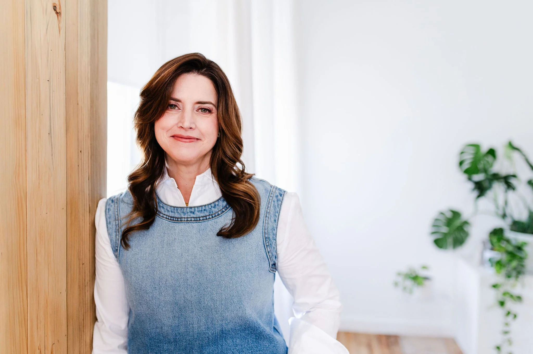 A woman with brown, shoulder-length hair, wearing a white shirt and a denim jumper, standing next to a wooden wall with plants in the background.