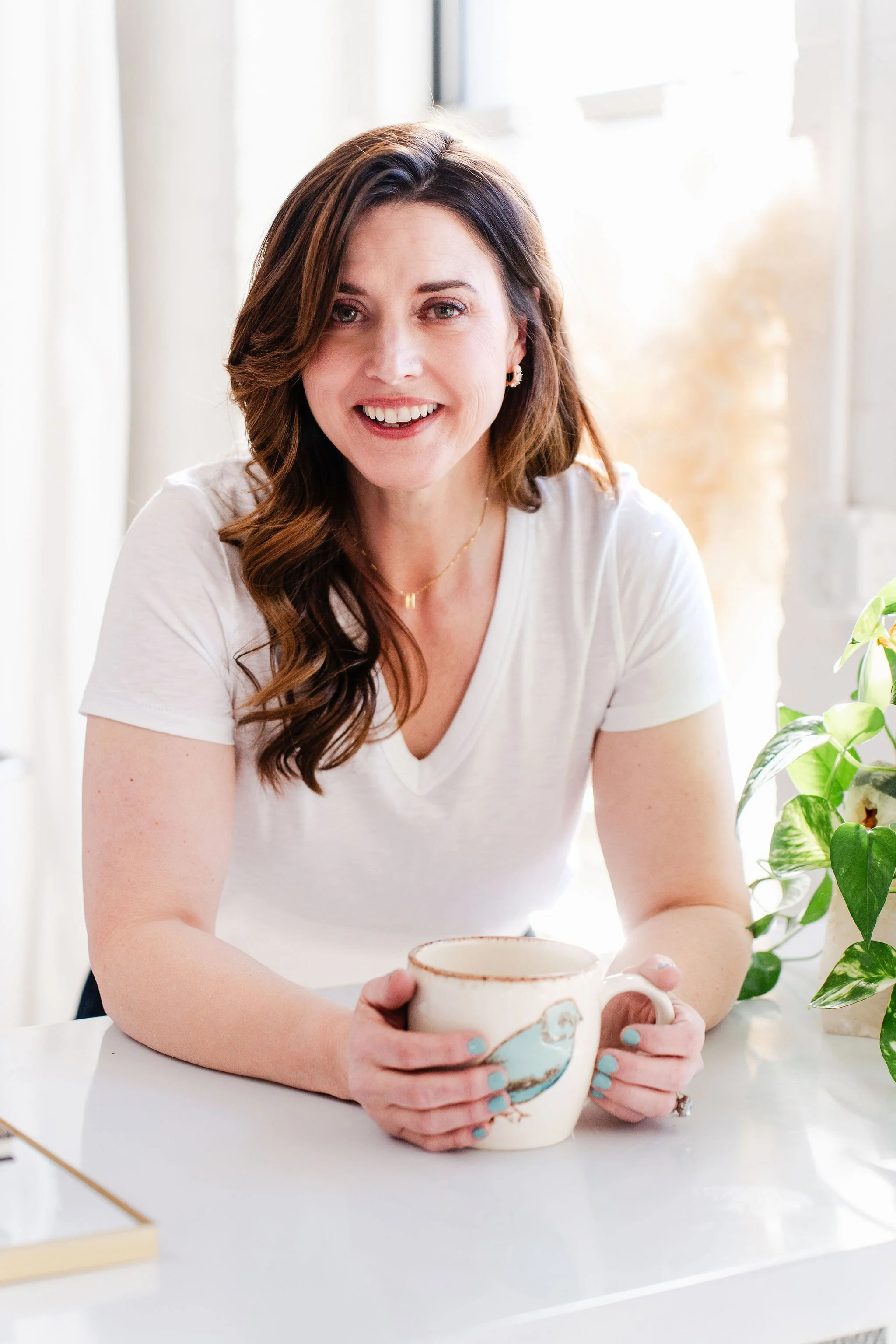A woman with shoulder-length brown hair holding a decorative ceramic mug with a floral design, sitting at a white table with a potted plant nearby, in a bright room with white curtains and windows.
