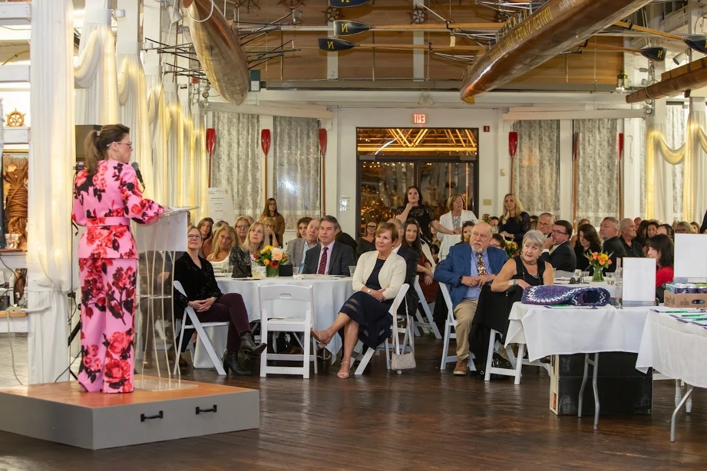 A woman in a floral pink suit speaking at a podium during a formal event or conference in a decorated banquet room with seated audience.