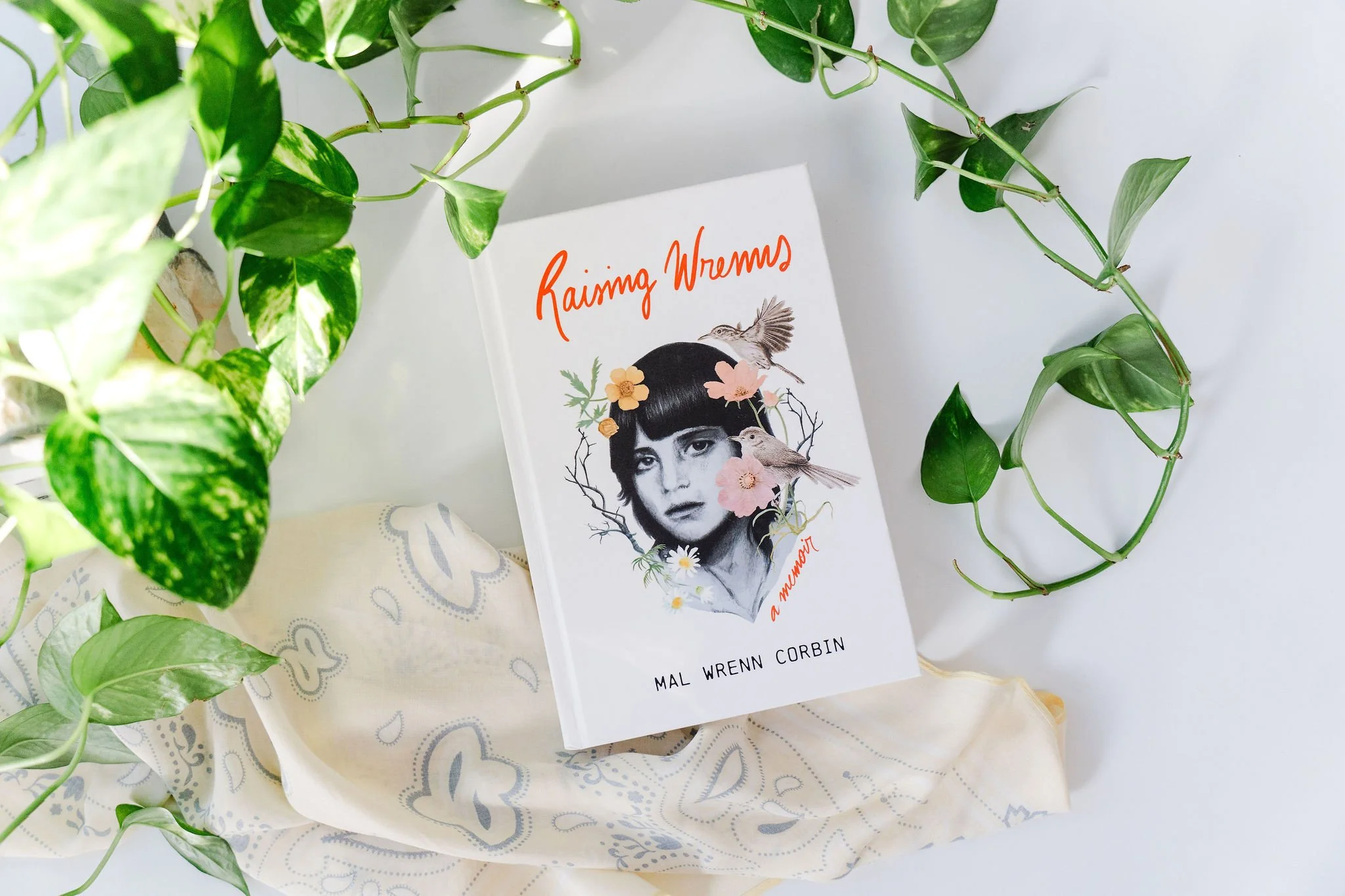 A book titled 'Raining Wrenns' by Mal Wrenn Corbin rests among green leafy plants. The book cover features a portrait of a woman's face with floral and bird illustrations.