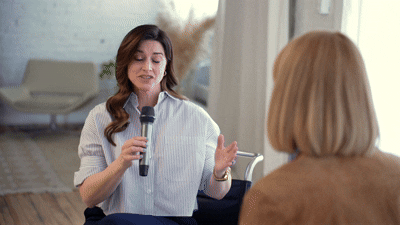 A woman with long brown hair speaking into a microphone during a discussion with another woman with blonde hair in a cozy indoor setting.