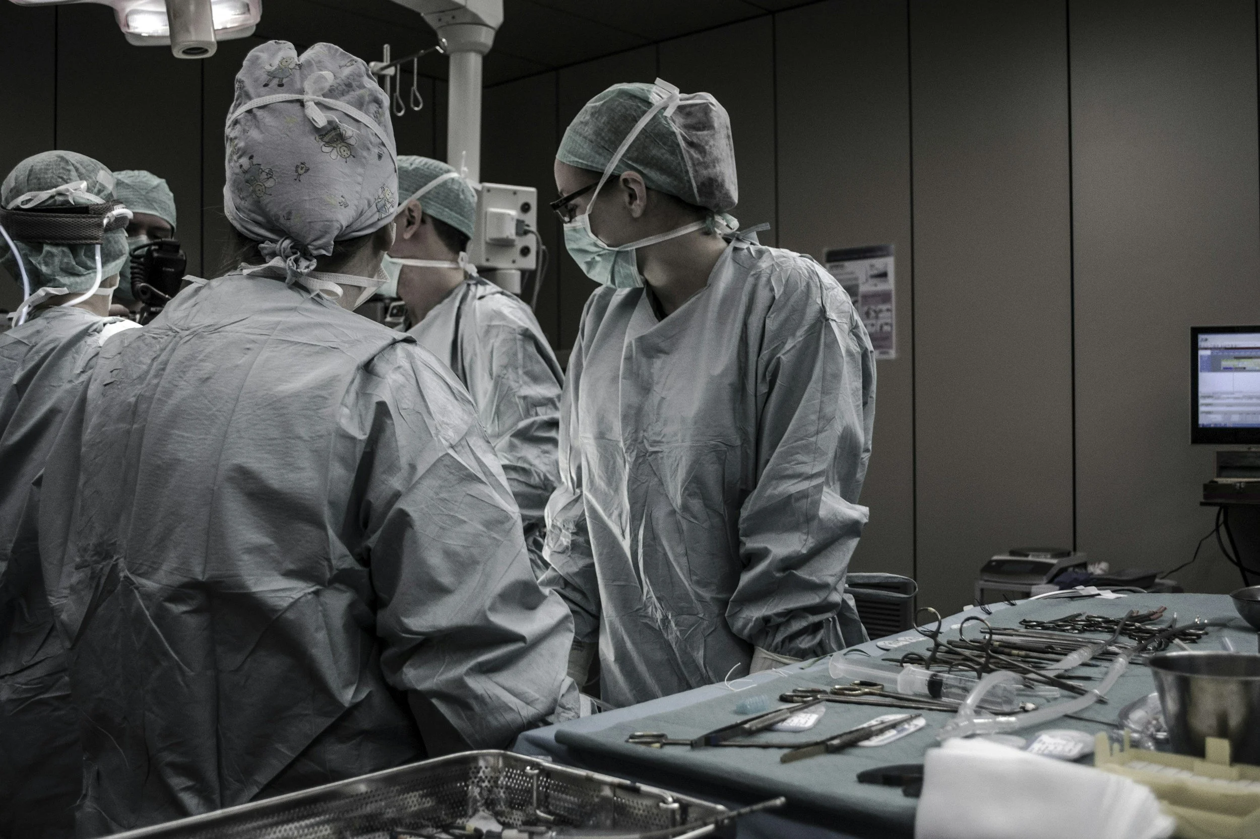 A team of surgeons in an operating room wearing scrubs, masks, and surgical caps, gathered around a patient for a procedure. Surgical instruments are laid out on a table.
