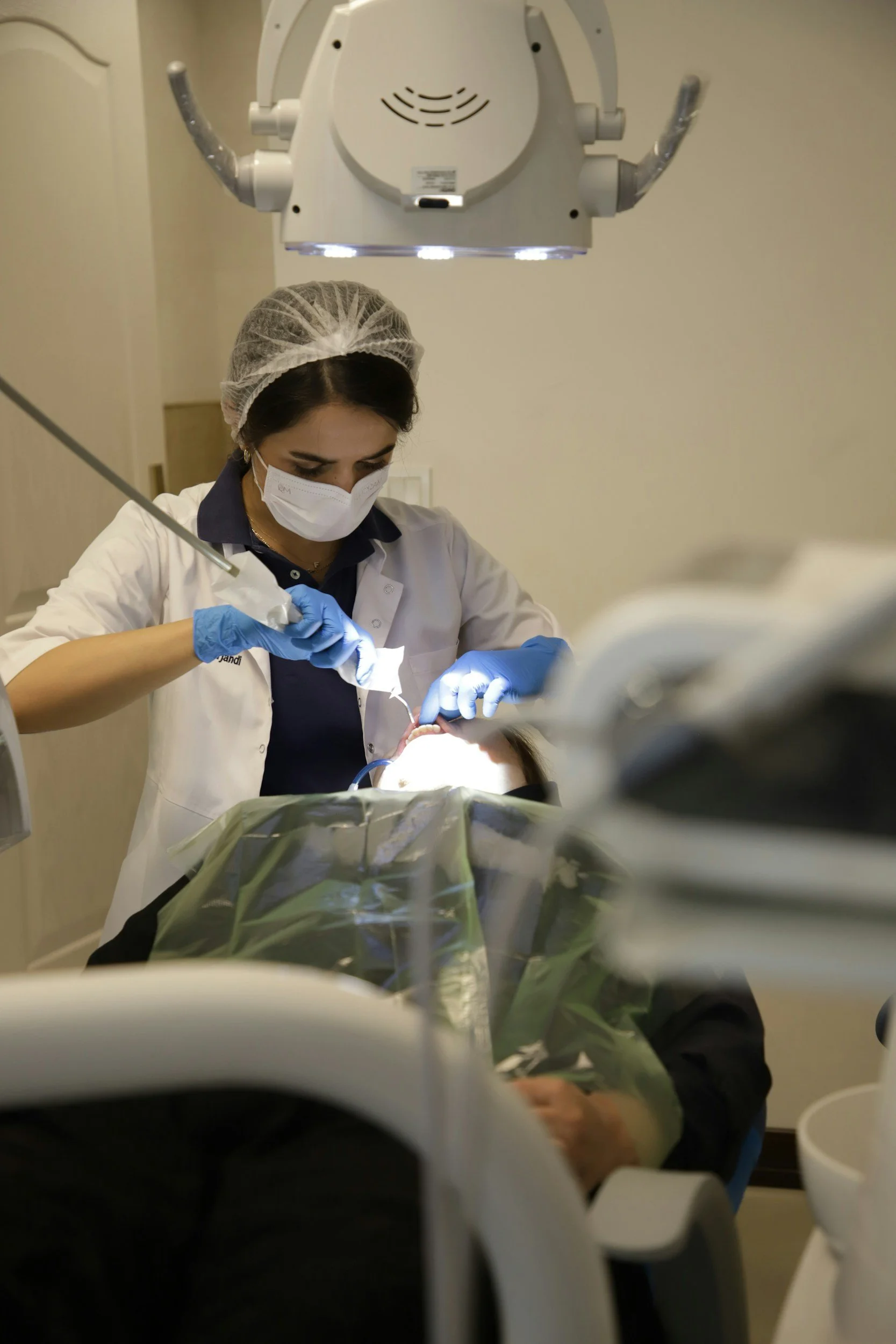 A healthcare worker performing a medical procedure, wearing a face mask, glove, and hairnet, beneath a surgical light in a clinical setting.