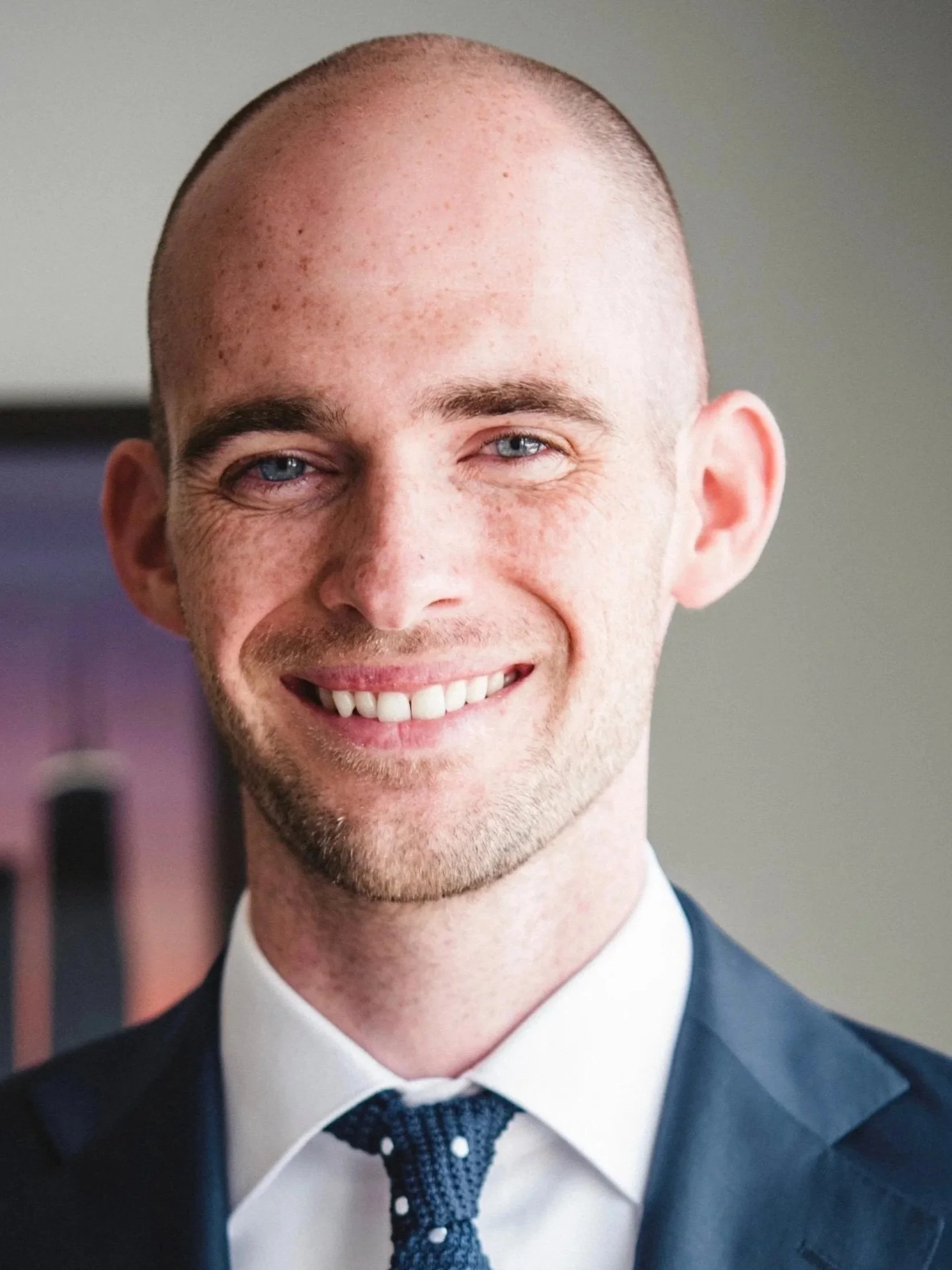 A smiling man in a suit with a white shirt and a navy blue tie featuring white polka dots.