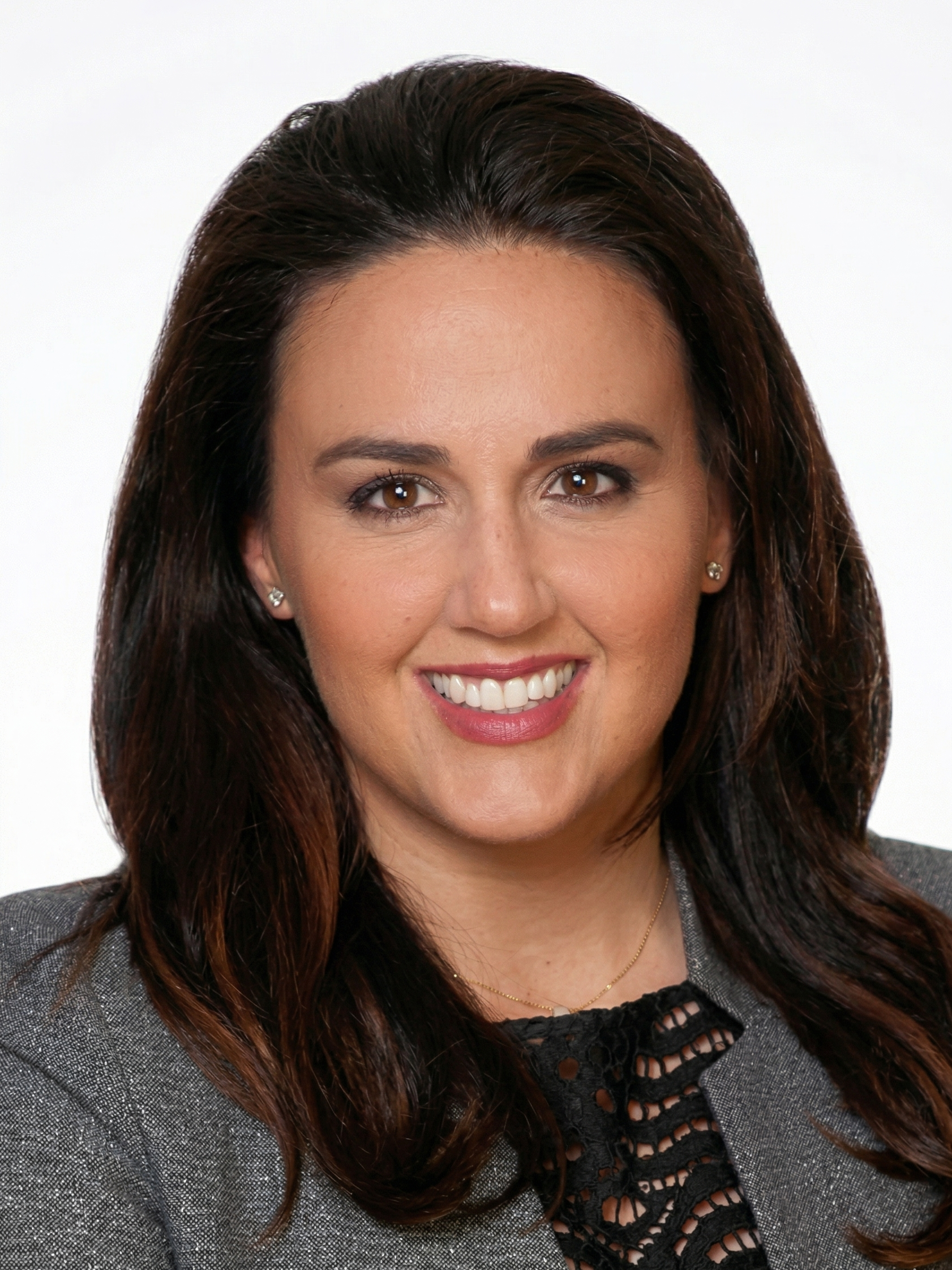 A professional woman smiling with brown eyes and long dark brown hair, wearing a grey blazer with black lacy top underneath, small earrings, and a delicate necklace.