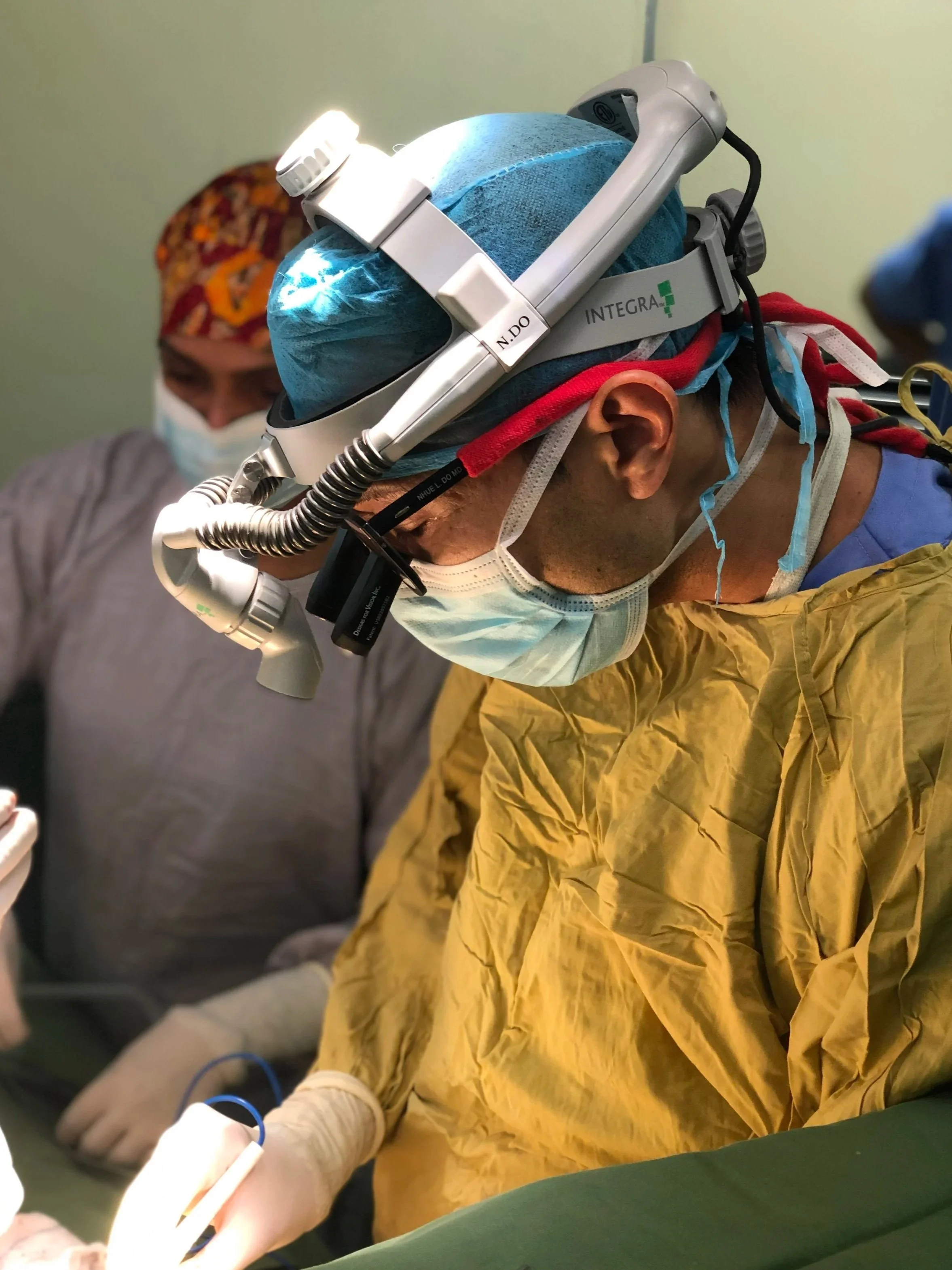 Cardiothoracic surgeon and real estate agent Nhue Do, wearing yellow gown, mask, and surgical cap, using surgical loupes and a headlamp during a medical procedure.