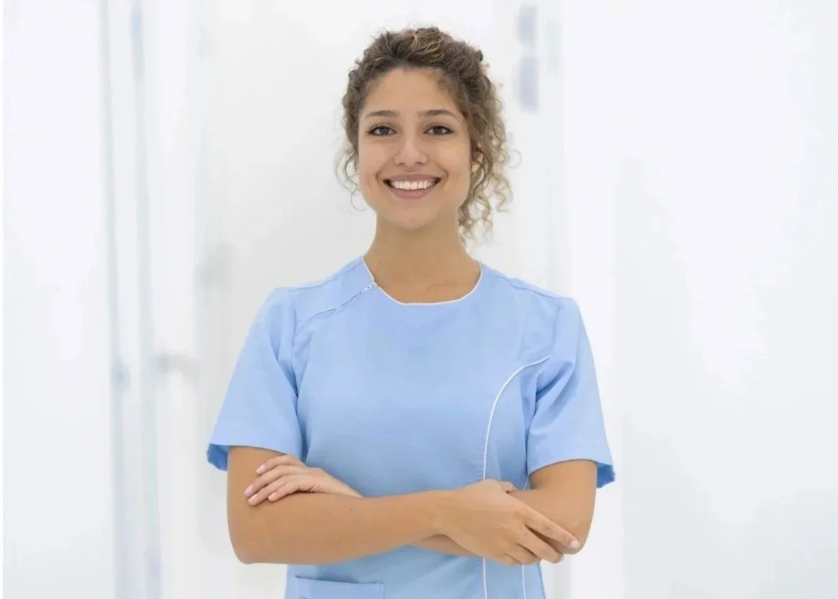 Young female nurse smiling, wearing light blue scrubs, standing with crossed arms in a bright hospital or medical facility.