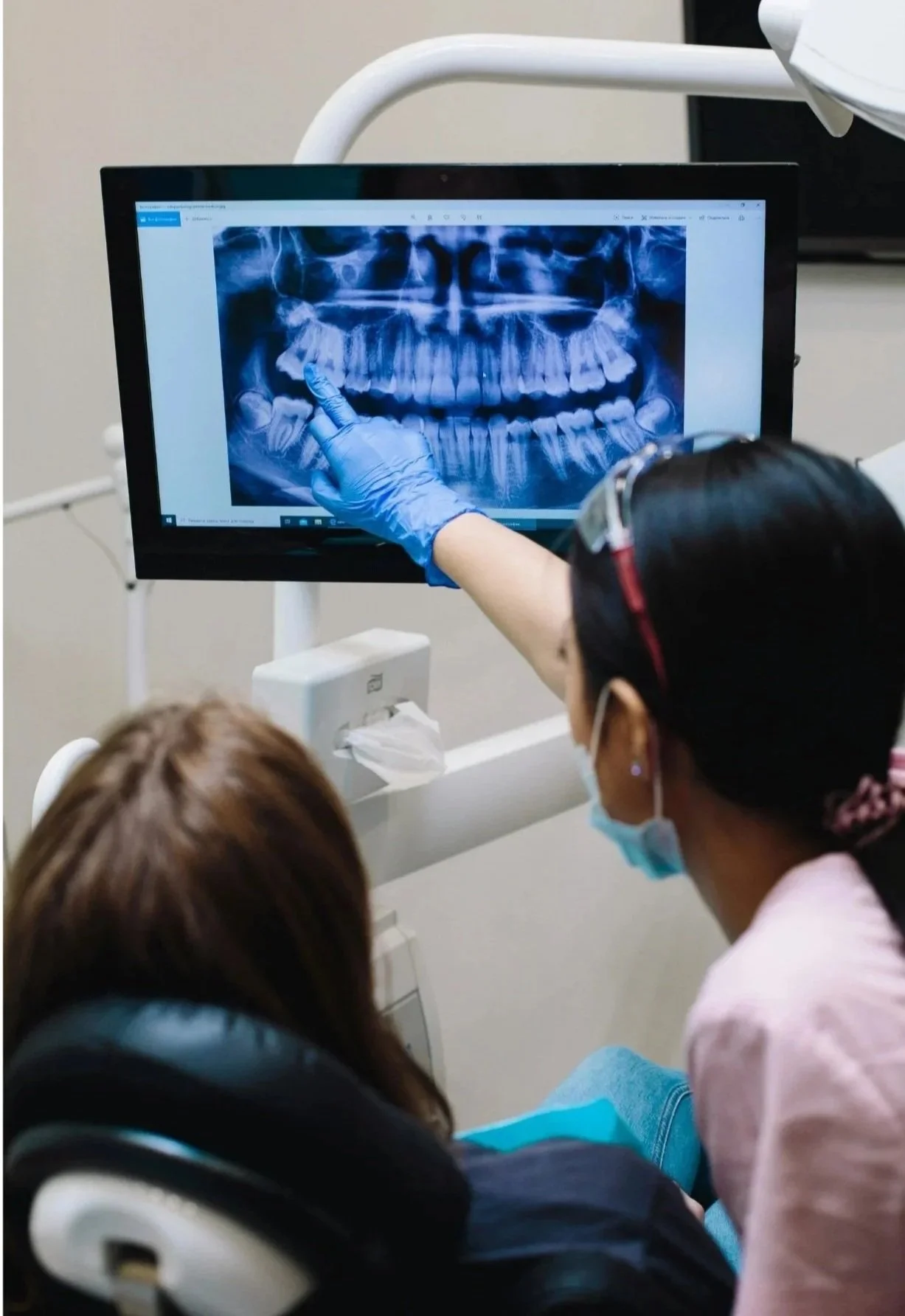 A dentist or dental assistant wearing gloves and a mask is pointing at a digital X-ray of a patient's teeth on a computer screen, with another person seated in front of the monitor, in a dental clinic.