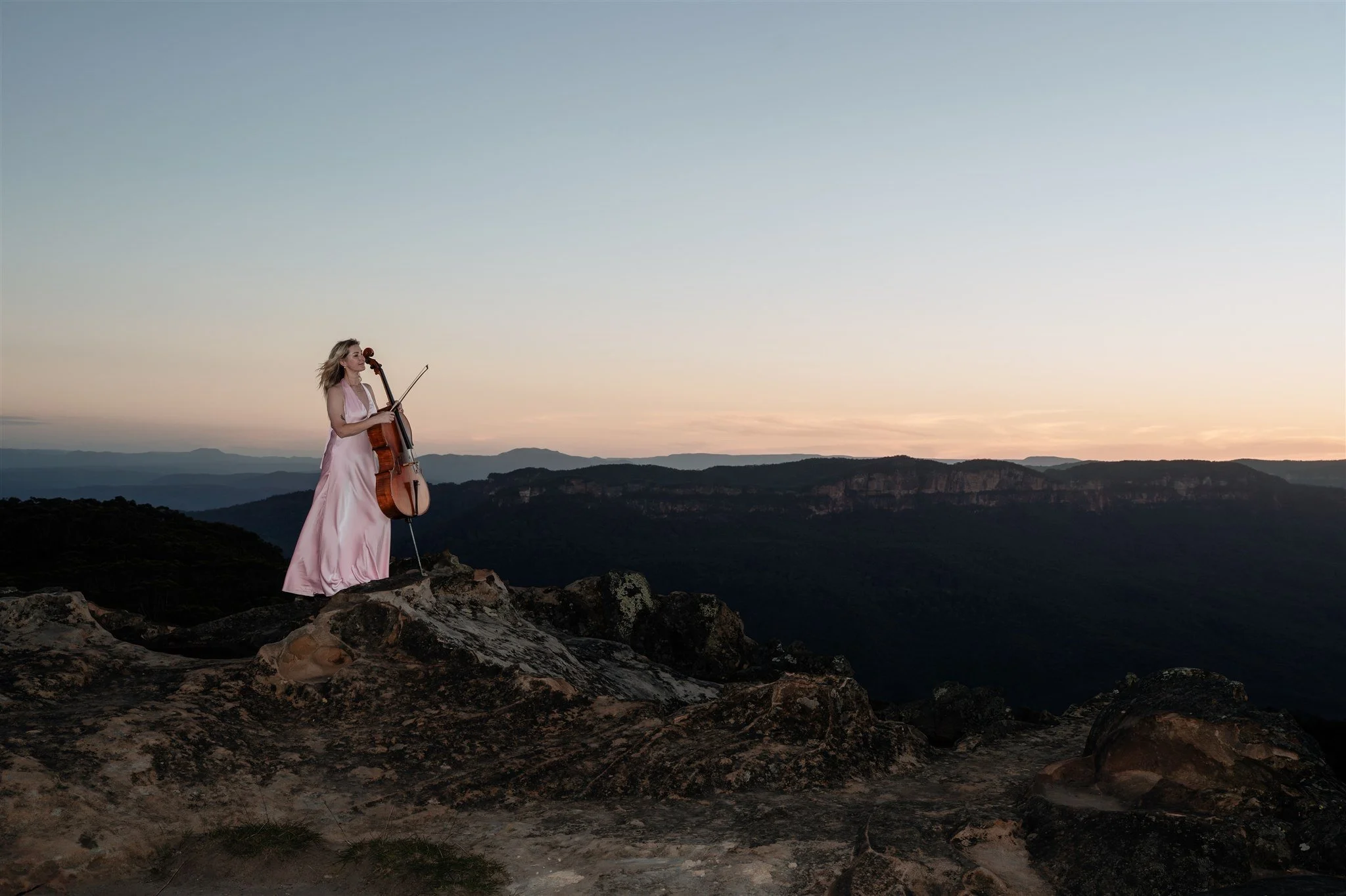 A woman in a pink dress is standing on a rocky cliff playing a cello at sunset, with mountains in the background.