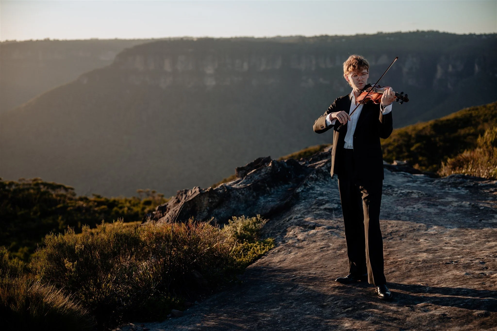 A man in a black suit and white shirt playing a violin outdoors on a rocky hill with mountains in the background at sunset.