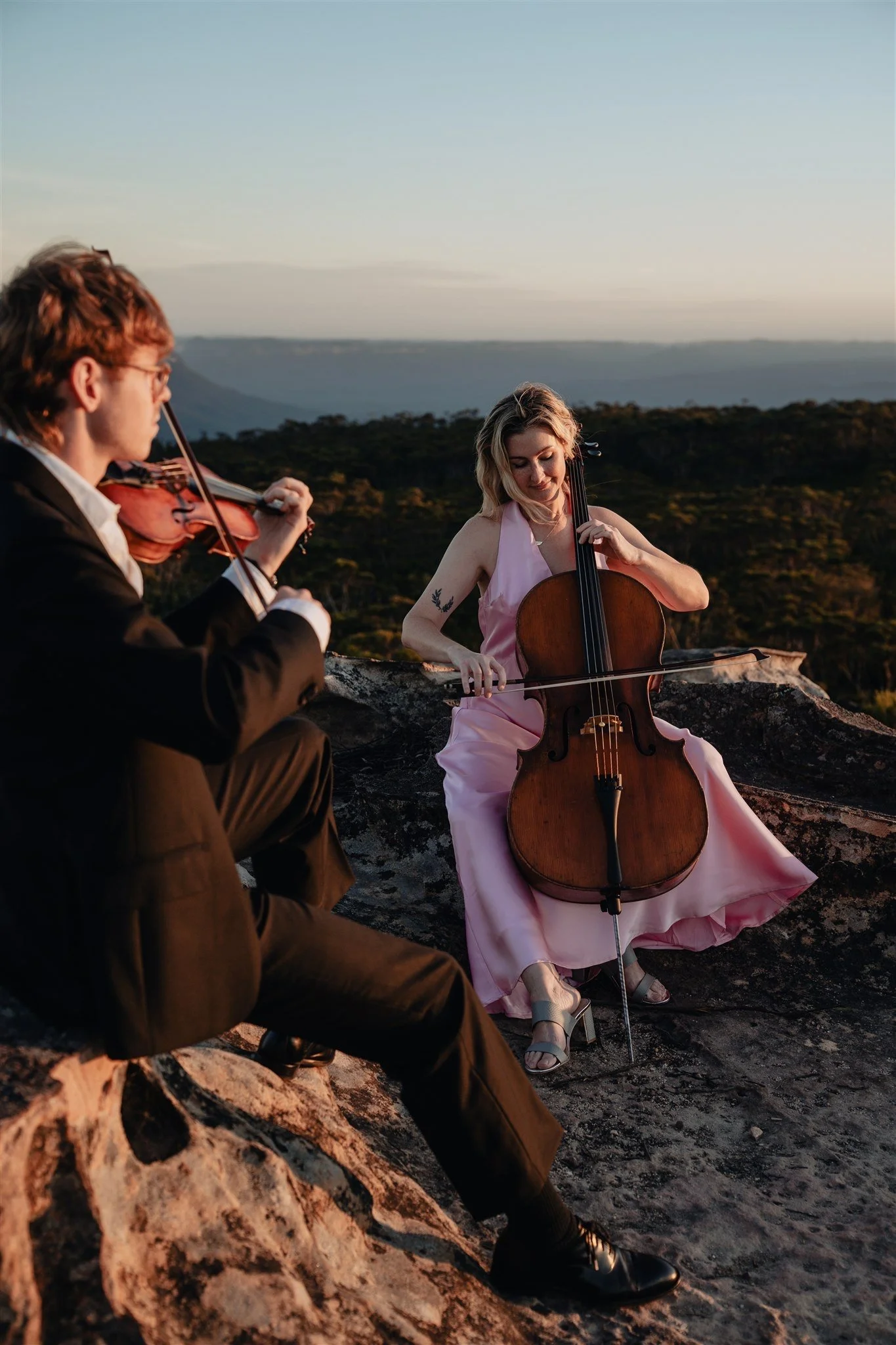 A woman in a pink dress playing the cello and a man in a suit playing the violin outdoors on a rocky surface during sunset.