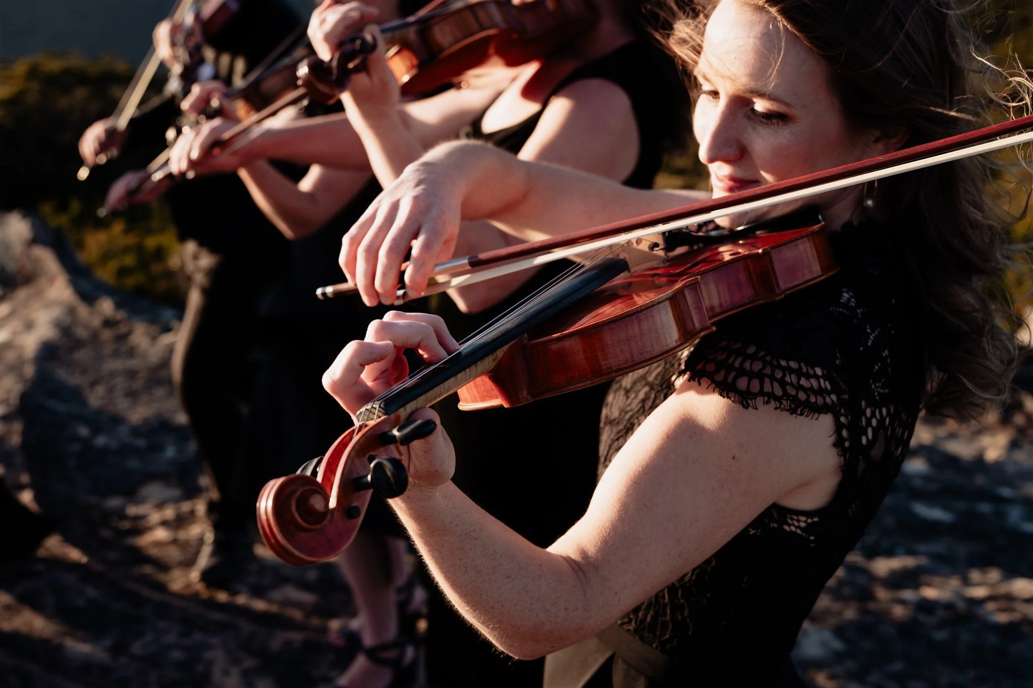 Group of women playing violins outdoors during sunset, focused and dressed in black.