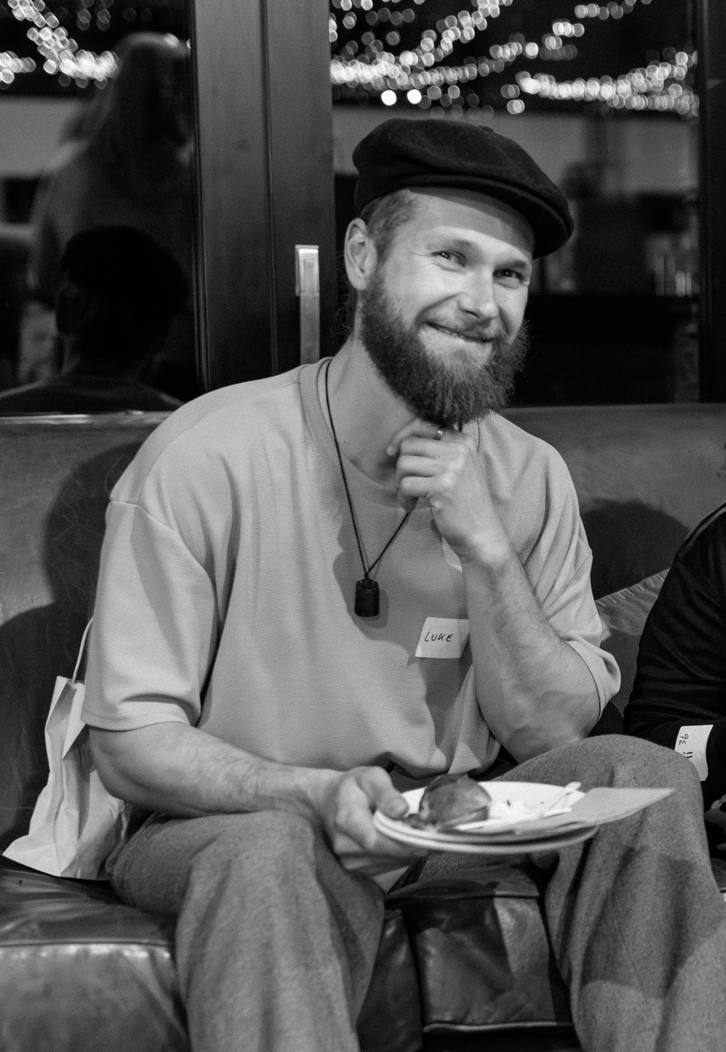 A smiling man with a beard, wearing a beret and a light-colored shirt, sitting on a couch in a dimly lit room, holding a plate with food, with string lights in the background.
