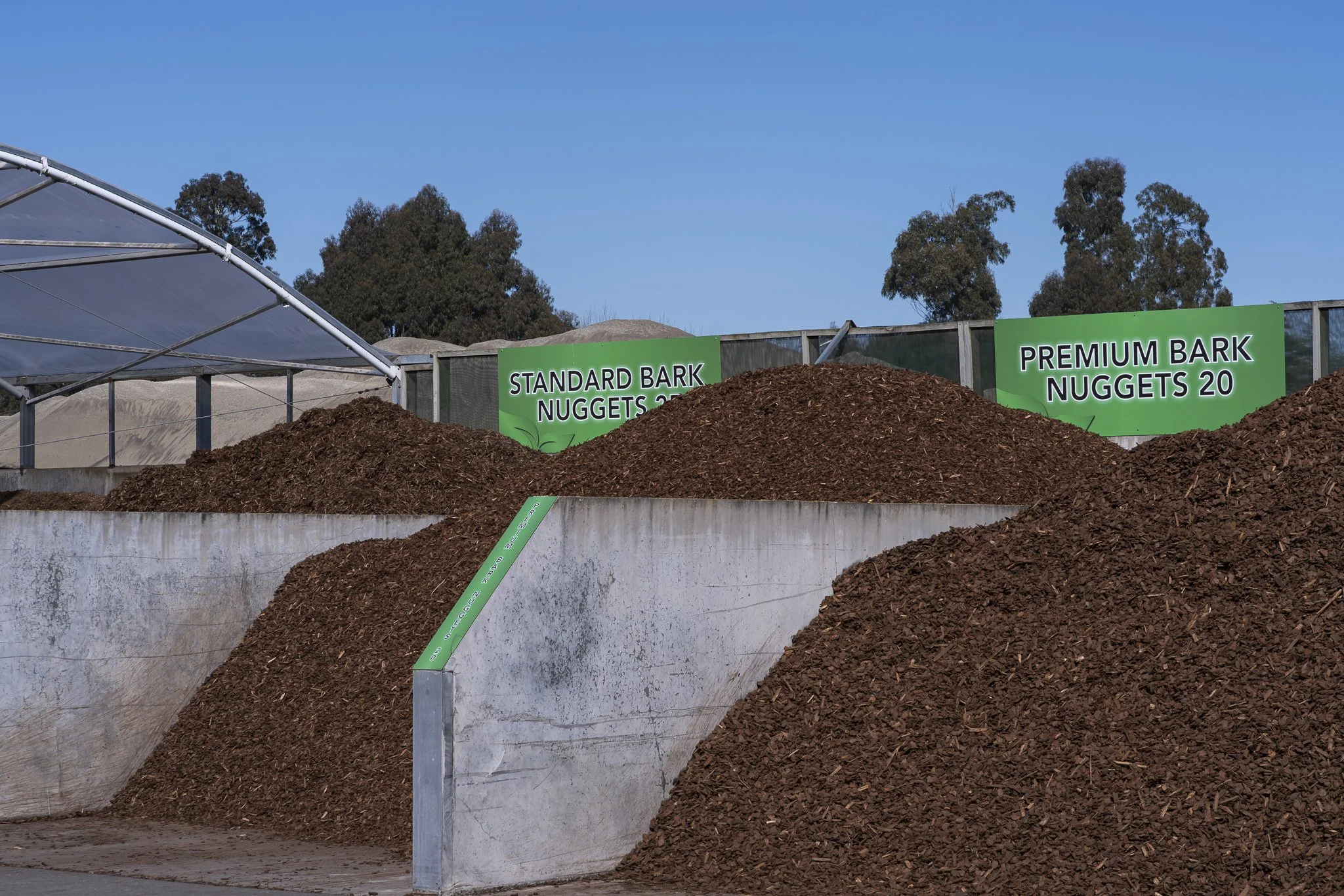 Display of mulch piles and signs indicating 'Standard Bark Nuggets 20' and 'Premium Bark Nuggets 20' at a landscaping supply yard with trees and a blue sky in the background.