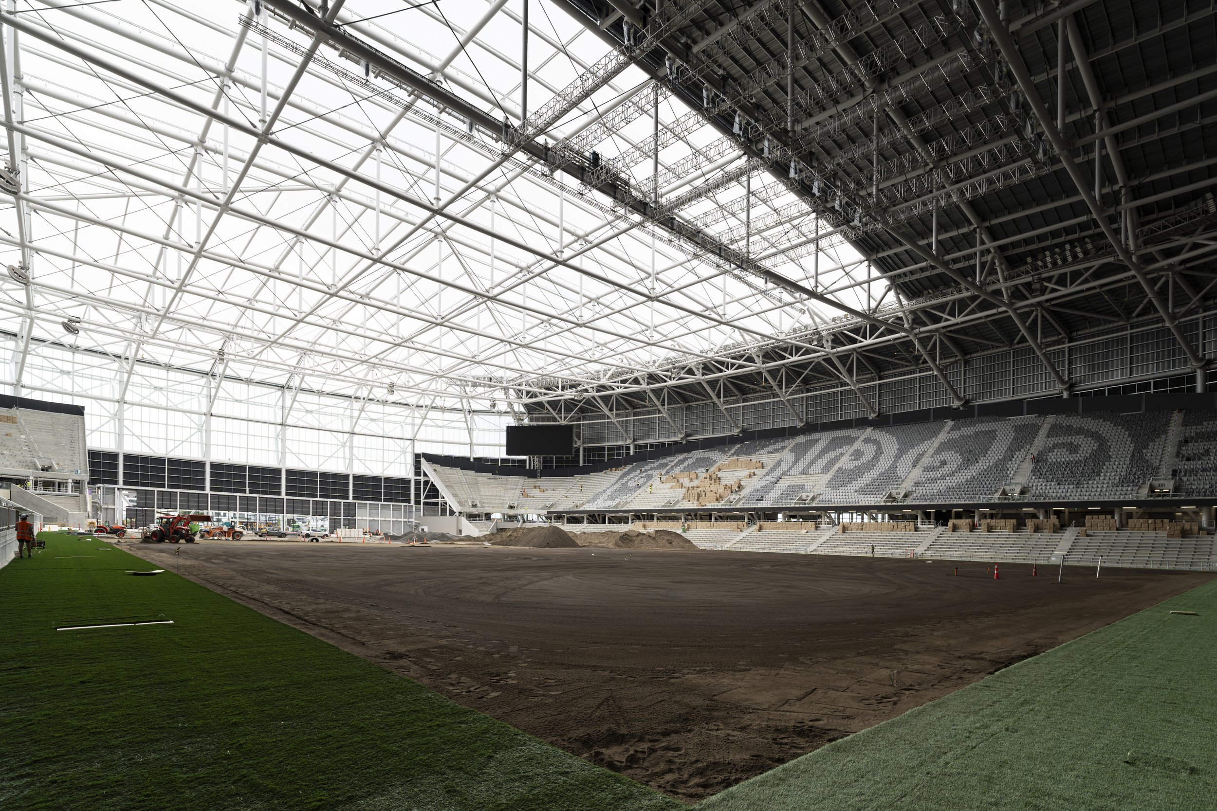 A stadium under construction with a large metal framework for the roof and seating areas. The field has artificial grass and construction equipment is present, indicating ongoing building work.
