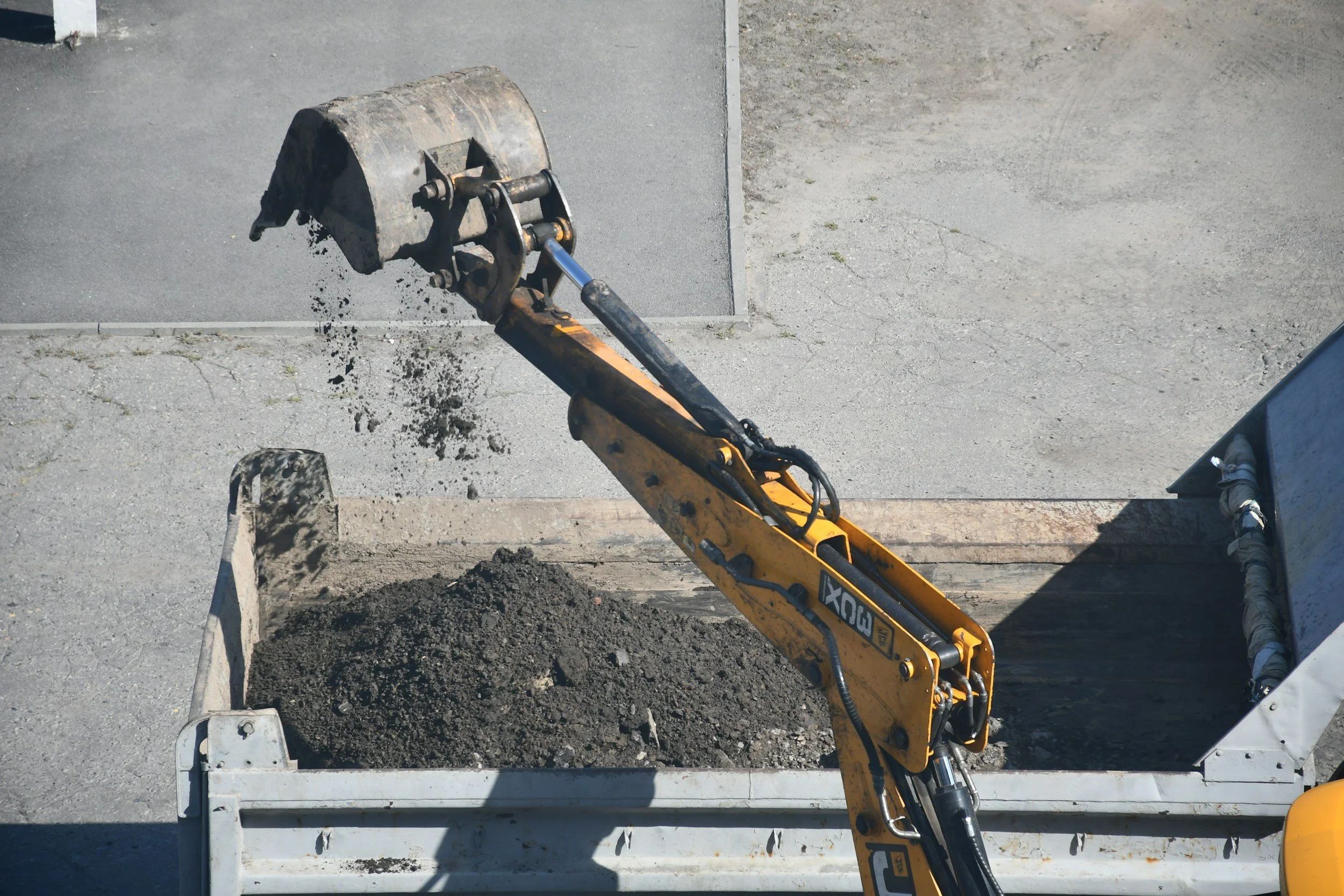 A yellow excavator with a bucket attachment digging into a pile of dirt inside a construction zone. The area has gray concrete and asphalt surfaces.