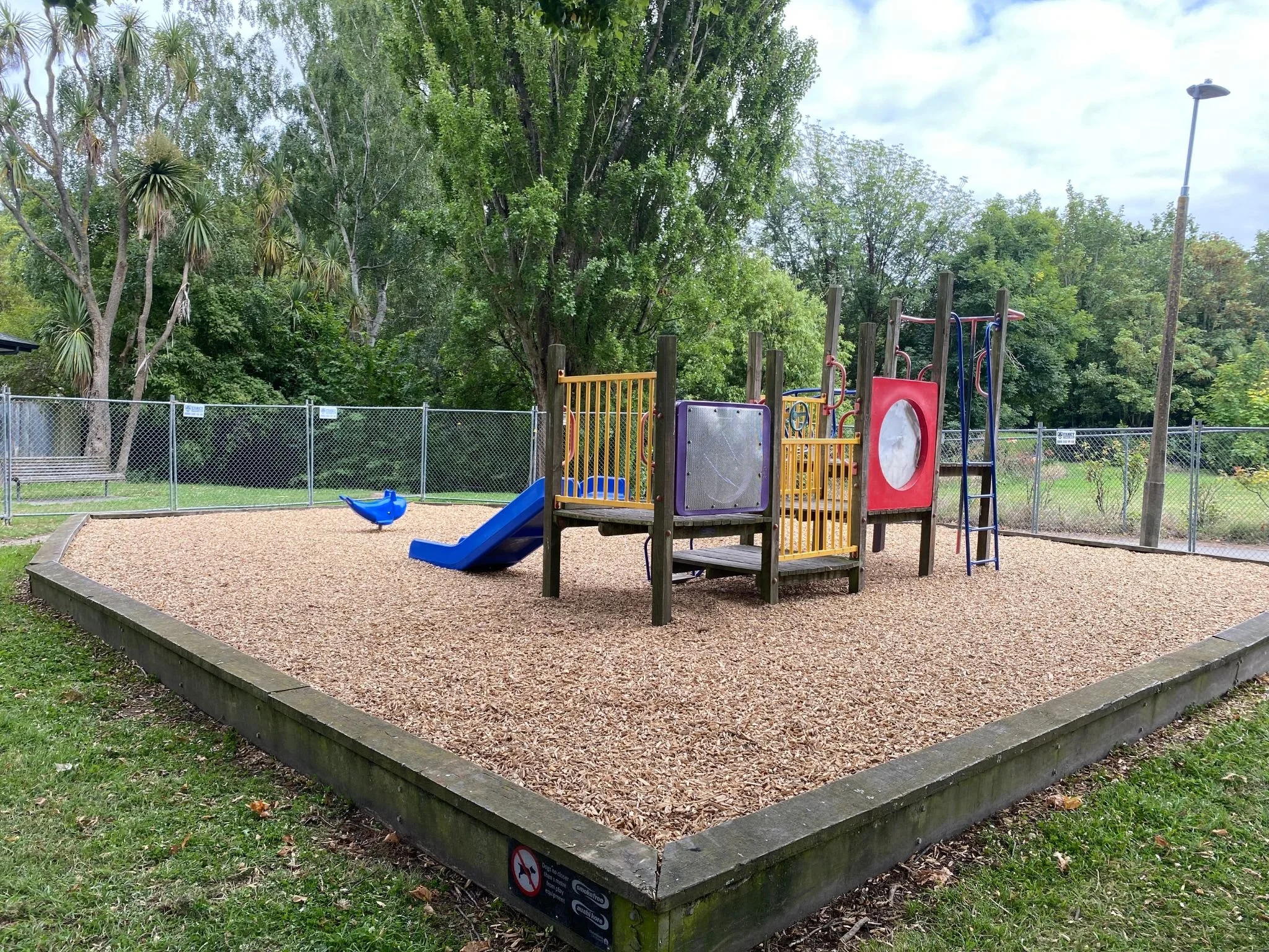 A playground with a blue slide, sand surface, surrounded by green trees and a fence. A streetlamp is visible in the background.