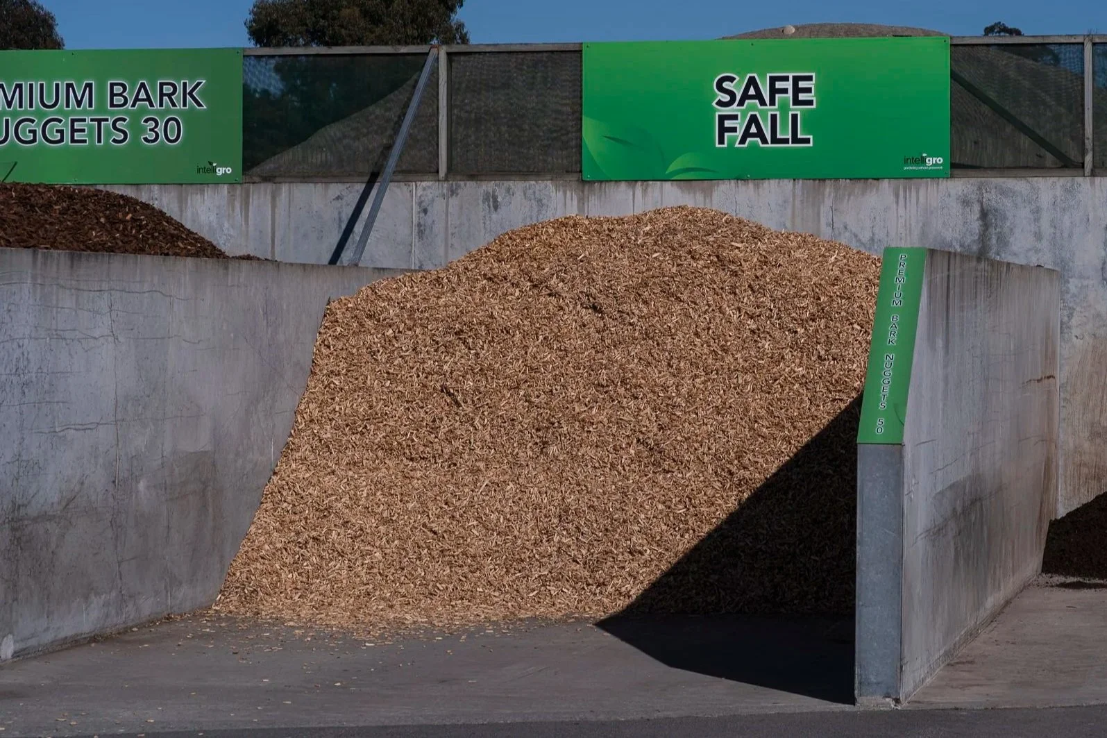 Pile of wood chips or mulch underneath green signs that say 'Premium Bark Nuggets 30' and 'Safe Fall', with a smaller green sign on the concrete wall indicating 'Premium Bark Nuggets 50'.