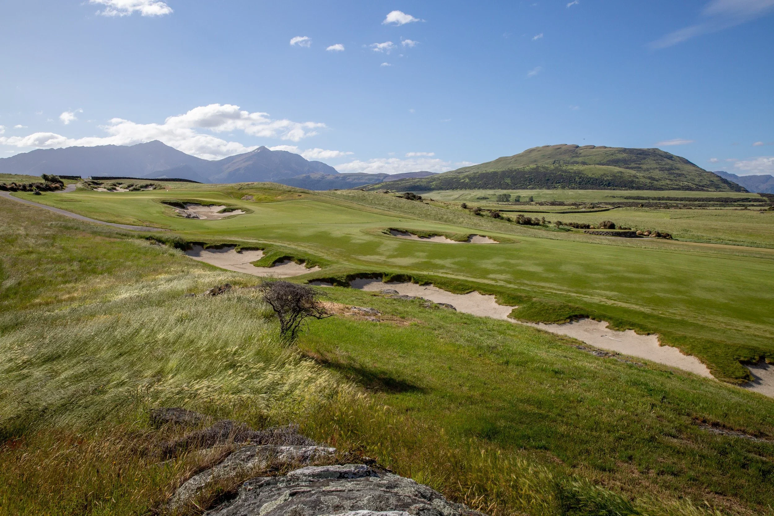 A scenic view of a golf course with green grass and sand traps, surrounded by rolling hills and mountains under a blue sky with scattered clouds.