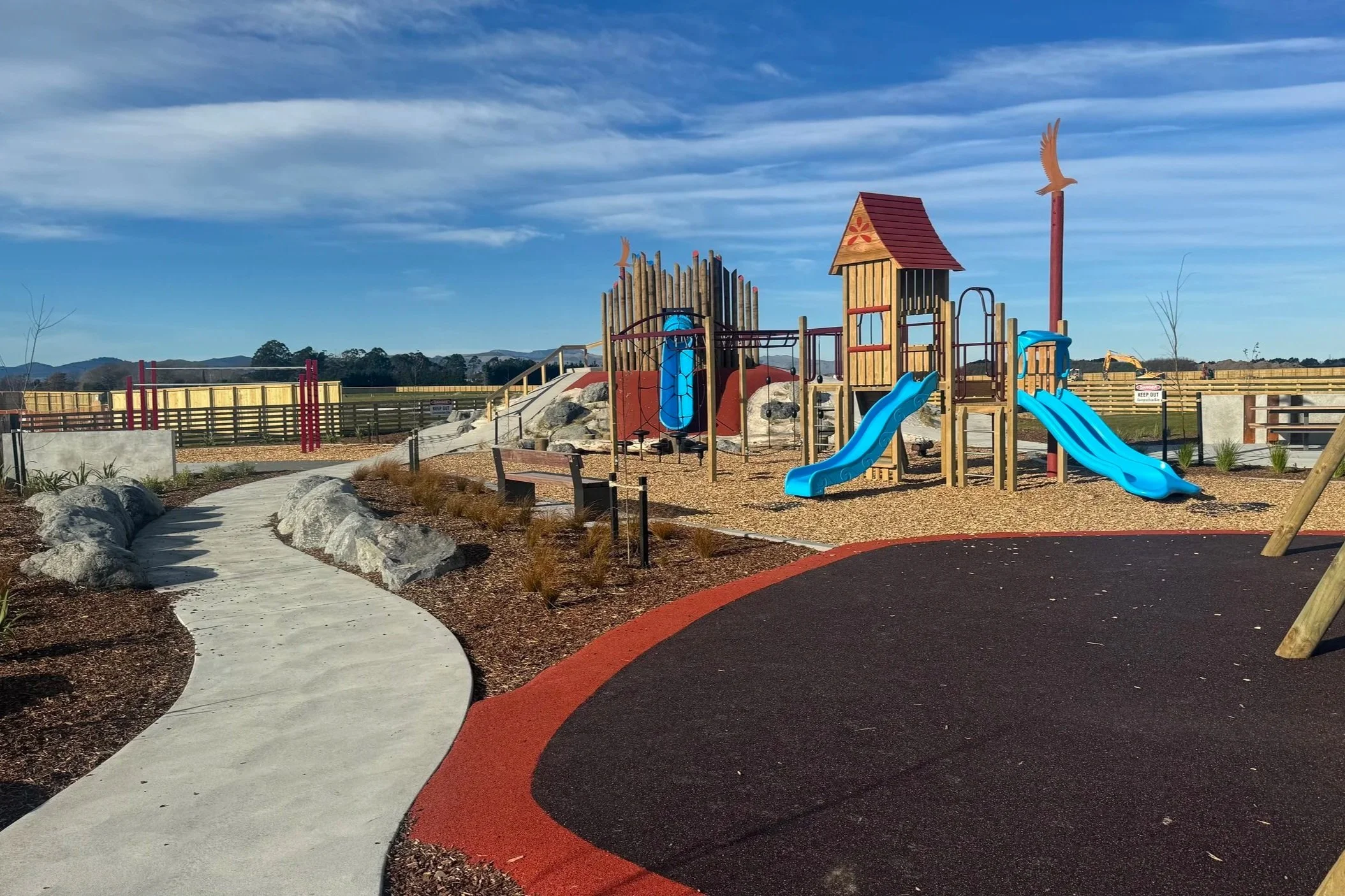 Children's playground with wooden structures, blue slides, and a slide shaped like a bird, surrounded by a fence and a mountain backdrop under partly cloudy sky.