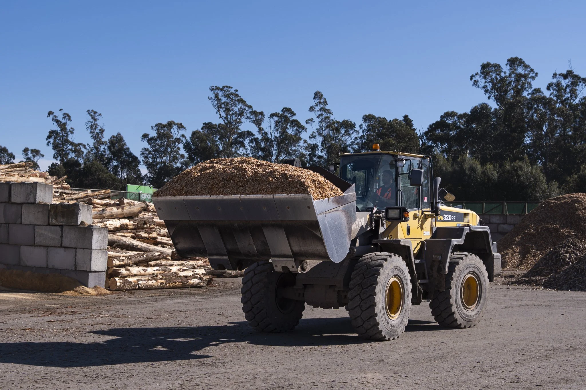 A construction site with a yellow front loader carrying a large pile of wood chips, with stacks of logs and bricks nearby, and a line of trees in the background under a clear blue sky.