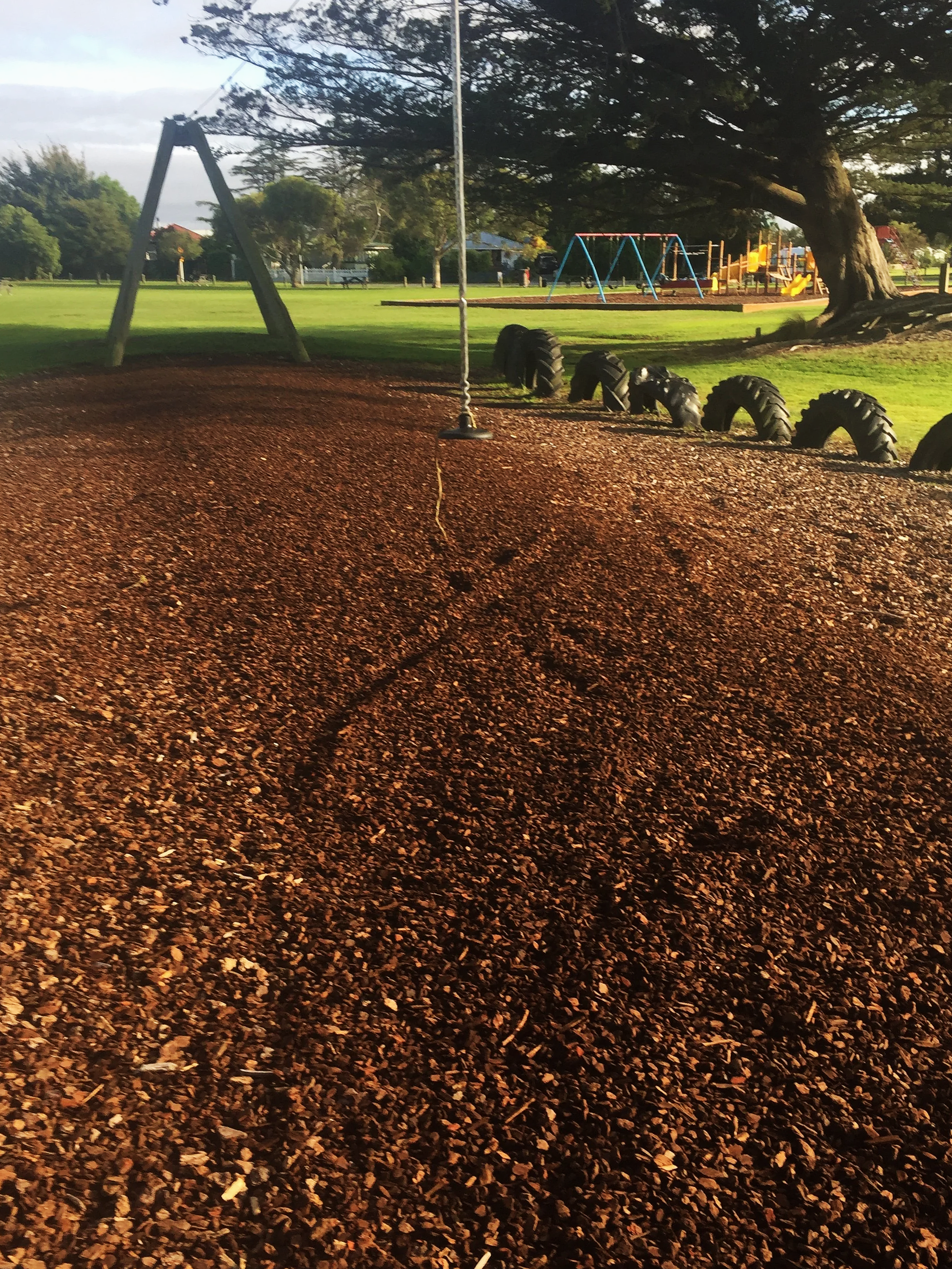 A playground with rubber mulch ground, a swing set, tires arranged on the ground, and a slide structure in the background, with a large tree providing shade.