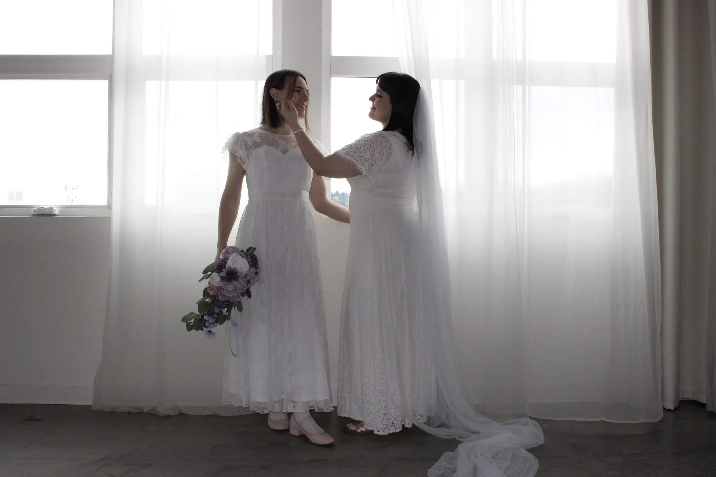 Two women in white wedding dresses standing inside by a large window with sheer curtains. One is holding a bouquet of purple and white flowers, and they are touching faces and smiling at each other.