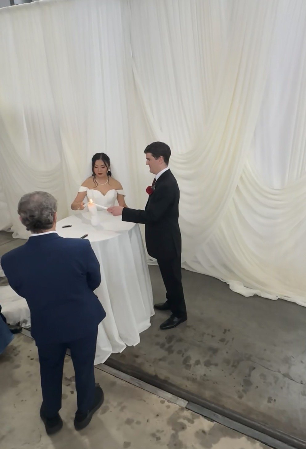 A wedding ceremony with a bride and groom lighting a unity candle, with an officiant observing, in front of a white draped backdrop.