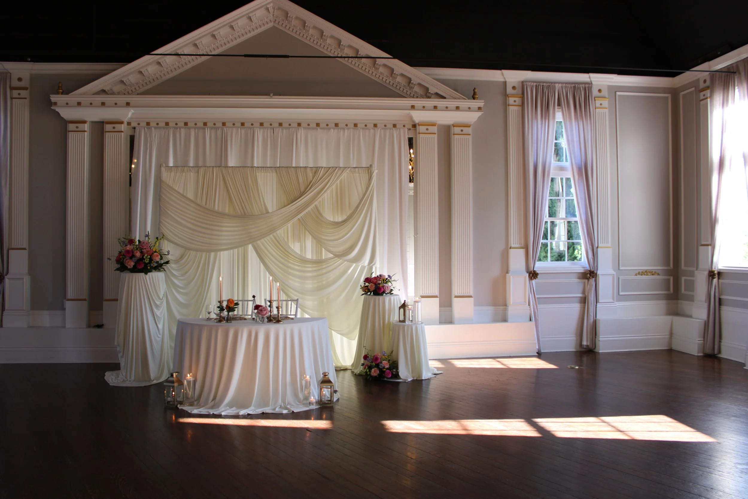 Elegant indoor wedding ceremony setup with draped white fabric, floral arrangements, candles, and chairs, in a room with white walls and tall windows with curtains.
