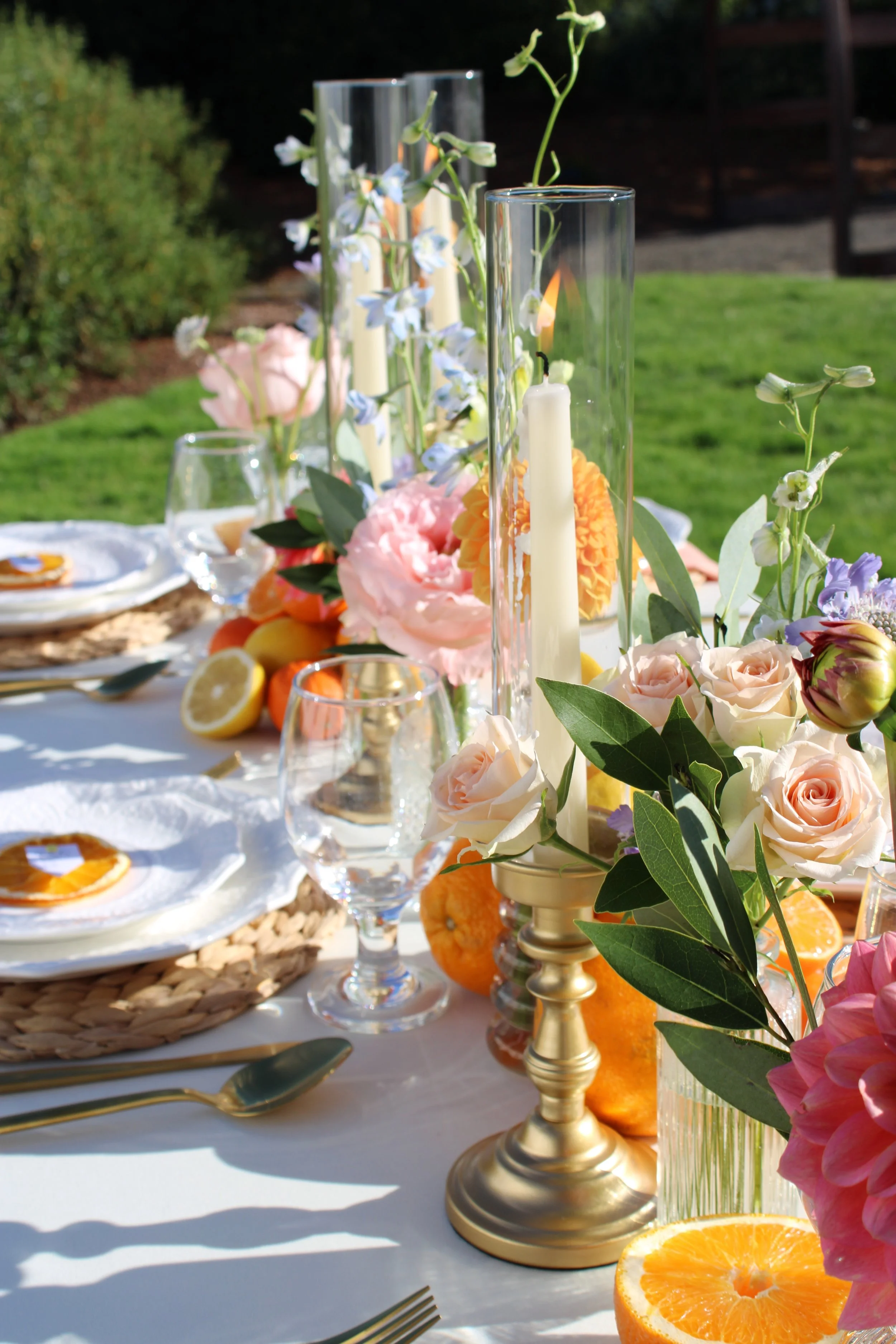 A festive outdoor dining table decorated with flowers, candles, and orange slices, set against a lush green yard with sunlight.