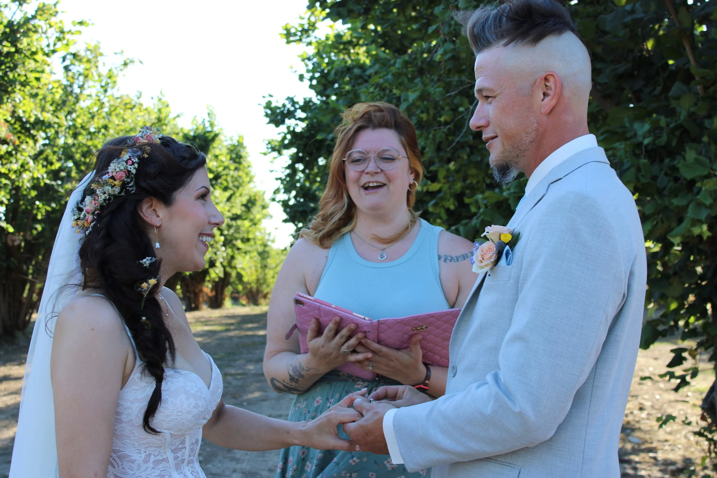 A couple getting married outdoors, holding hands, with an officiant standing between them, reading from a pink folder. The bride has dark hair adorned with flowers and wears a white lace dress. The groom has light hair with a mohawk and gray beard, d