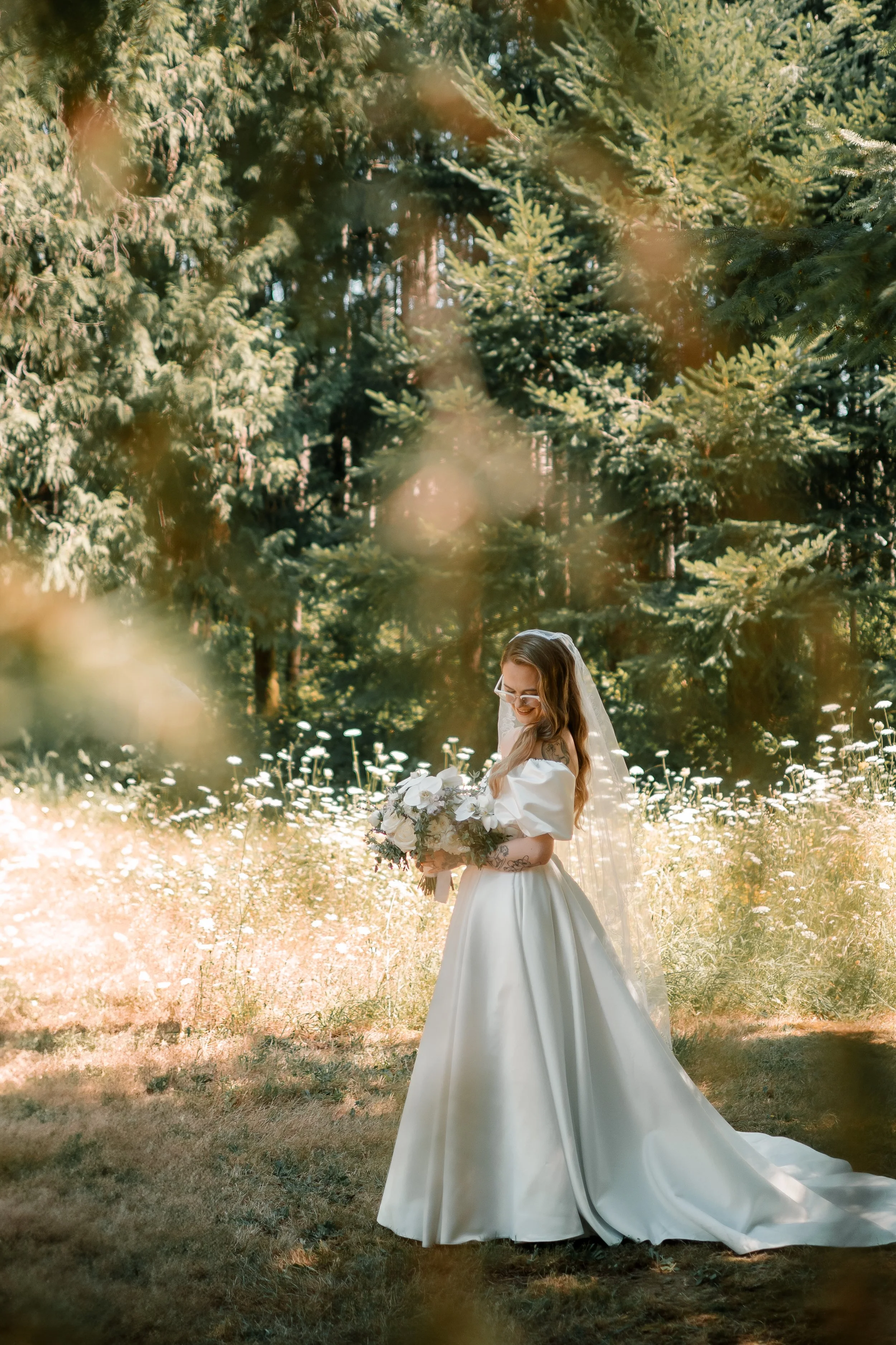 A bride in a white wedding gown holding a bouquet of flowers, standing in a sunlit forest with tall green trees and wildflowers.