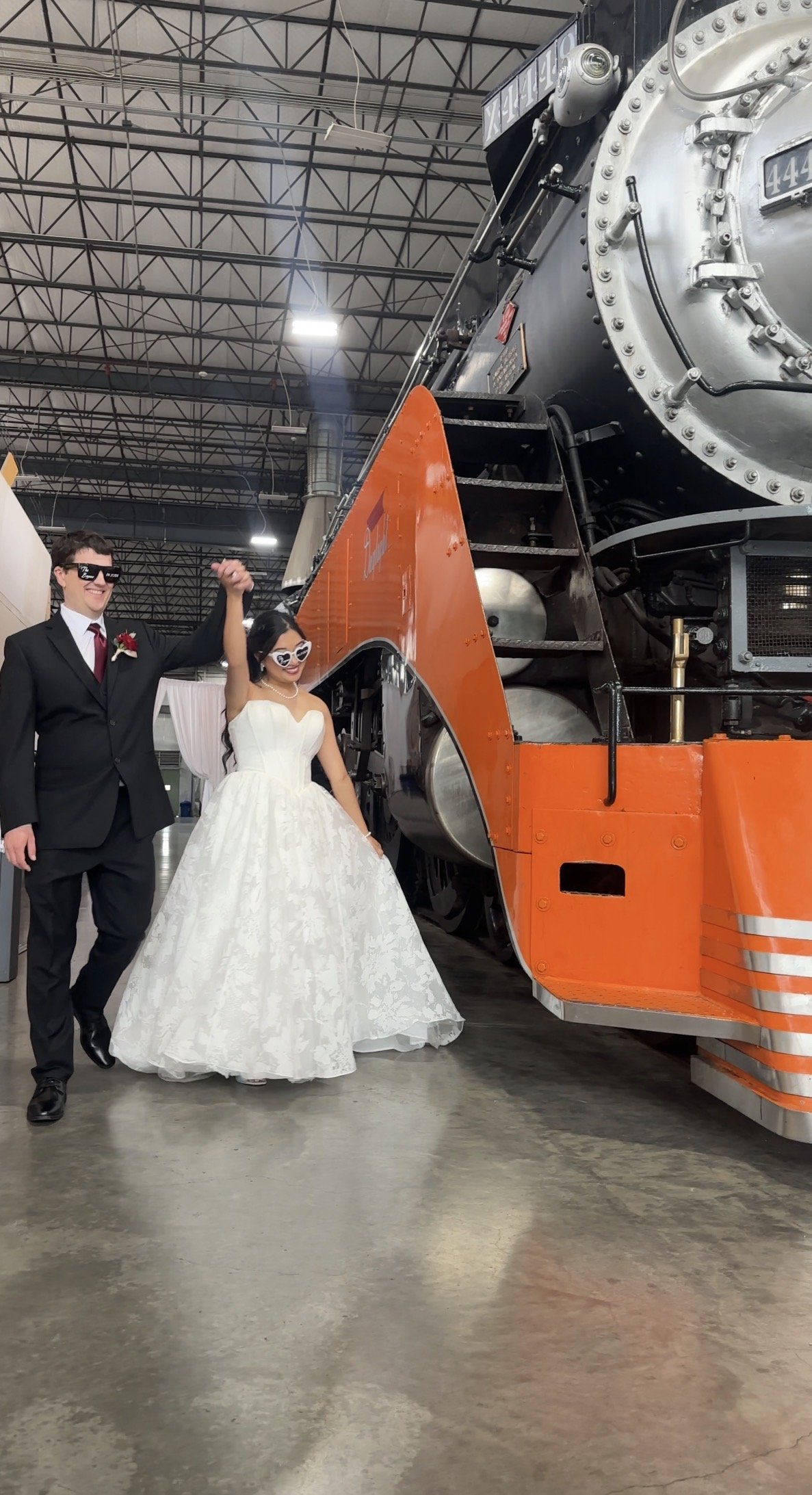 A bride and groom celebrating, standing next to a large vintage steam locomotive inside a spacious industrial-style building. The groom is wearing a black suit with a red tie, and the bride is wearing a white strapless wedding gown and sunglasses. Th