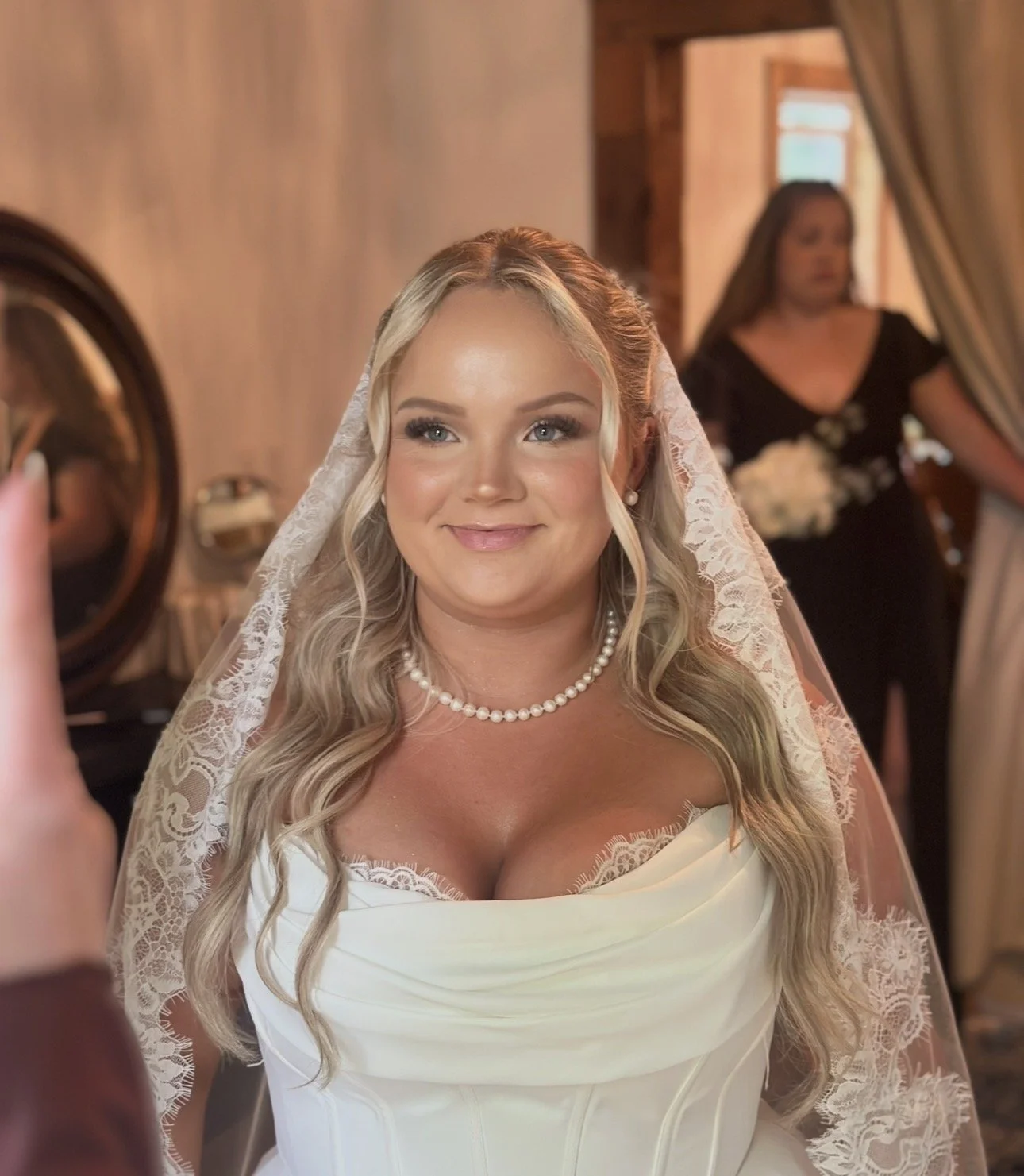 A bride with blonde hair, wearing pearl earrings and necklace, smiling as she gets ready for her wedding, with a woman in black behind her holding a bouquet, in a warmly lit room.