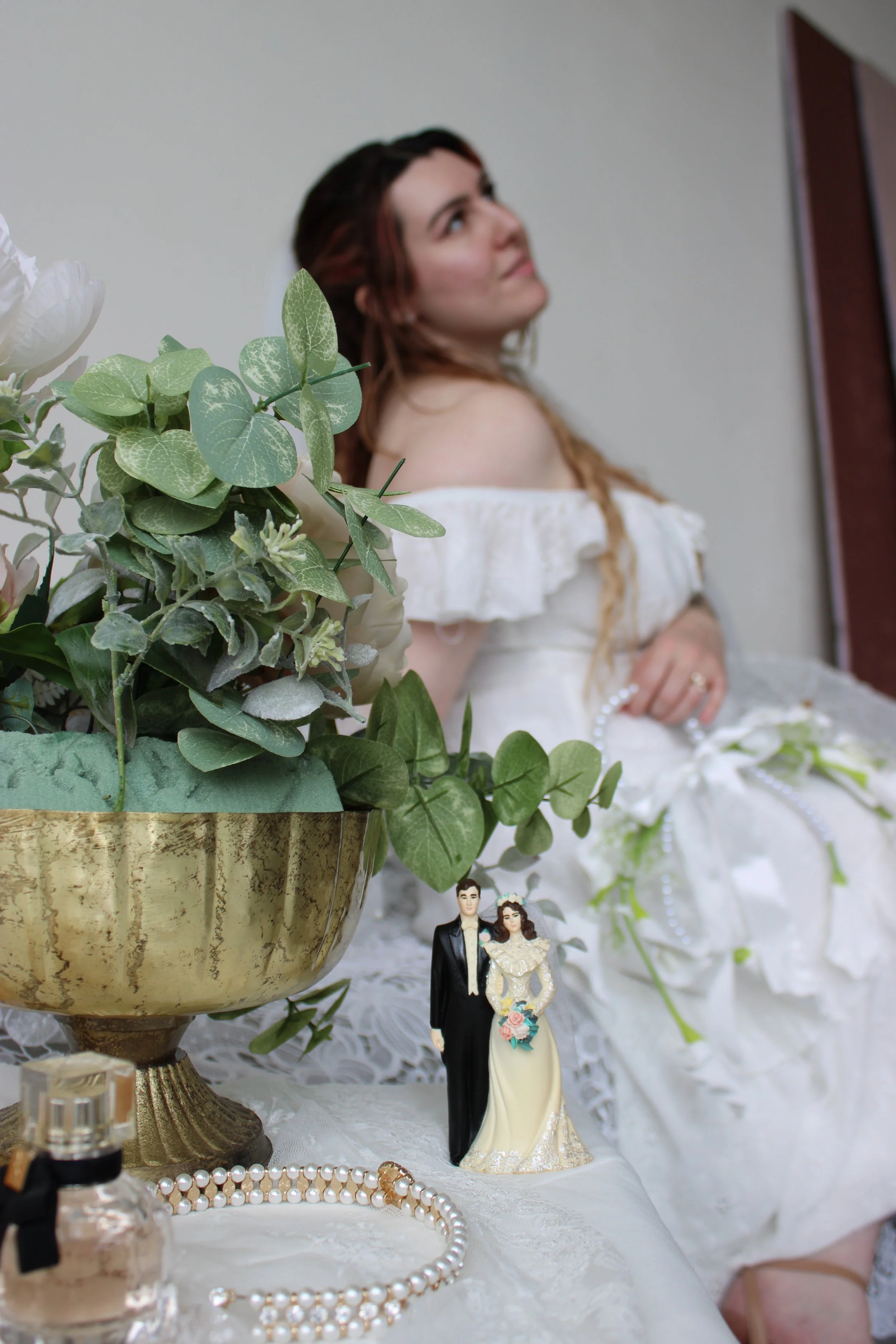 A bride in a white off-shoulder dress with lace details stands in the background, slightly out of focus. In the foreground, there is a large ornate gold-colored vase filled with green leaves and white flowers. A small figurine of a bride and groom, dressed in wedding attire, is positioned near the vase, surrounded by pearl jewelry and a small perfume bottle on a white lace tablecloth.