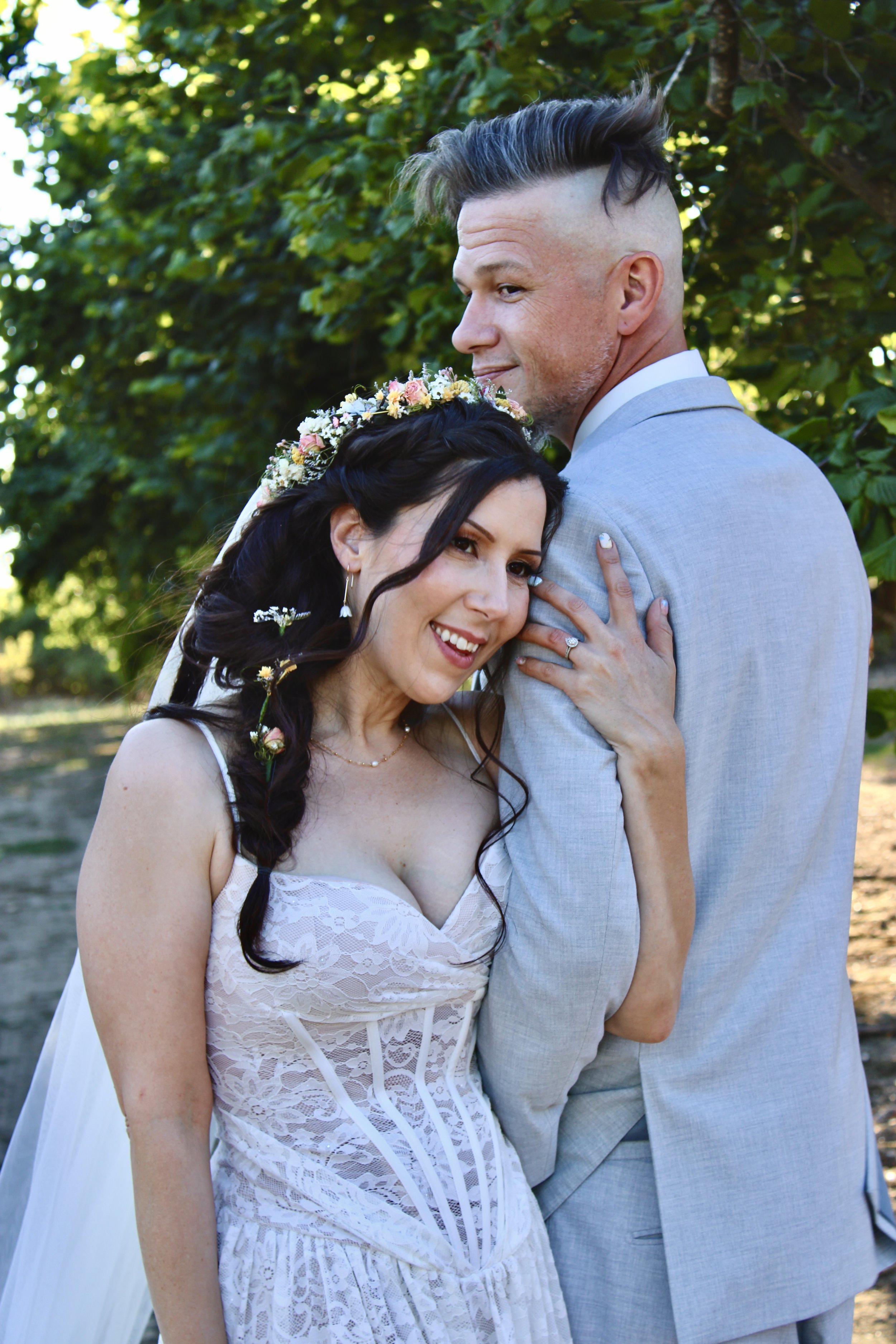 A bride and groom posing outside among green trees. The bride is smiling with her head resting on the groom's chest, wearing a white lace dress, a floral crown, and flowers in her hair. The groom is wearing a light gray suit, standing behind the brid