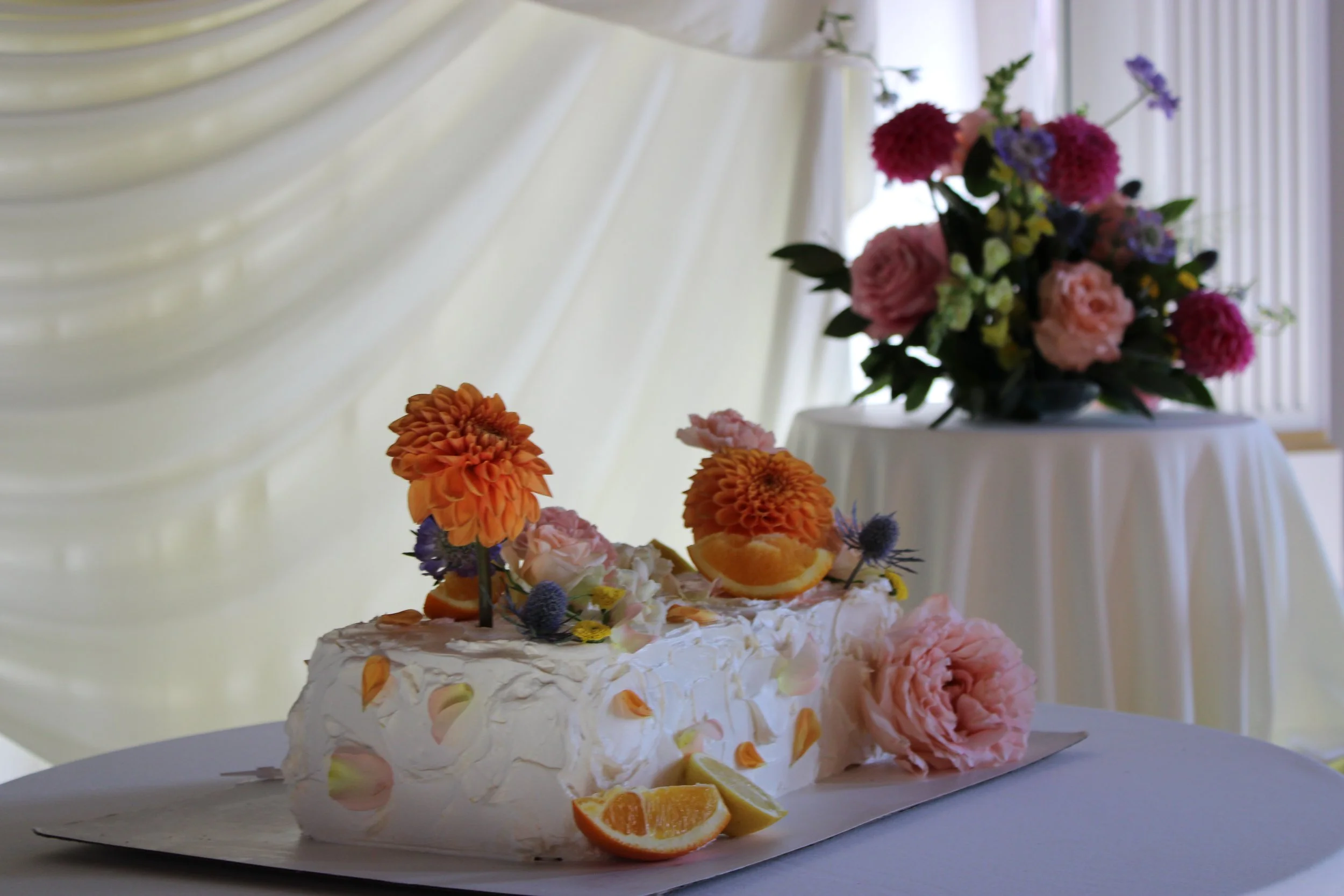 A decorated cake topped with pink, orange, and purple flowers, orange slices, and greenery, placed on a white table with a white draped backdrop and a large floral arrangement in the background.