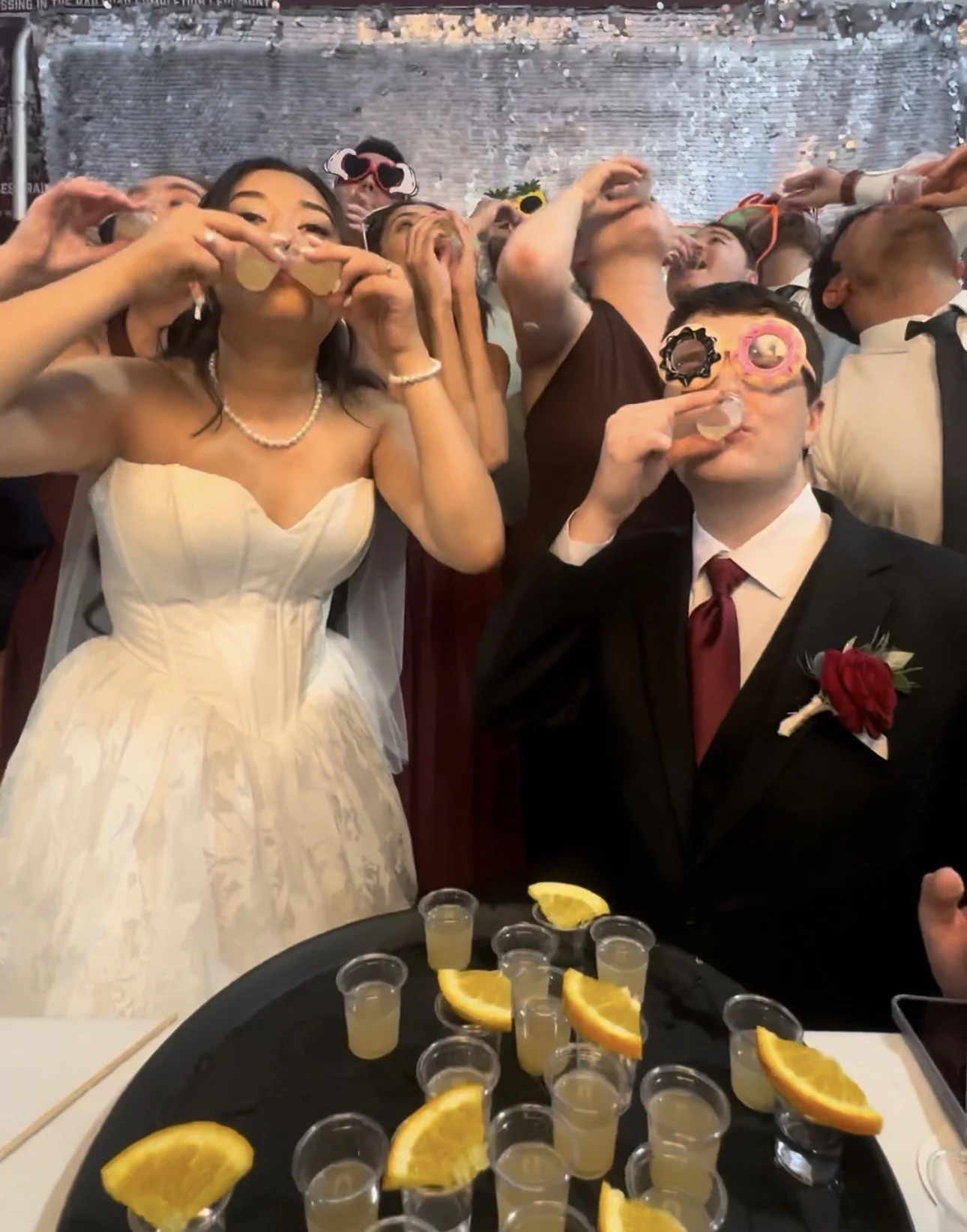 A crowd at a celebration, with people wearing party glasses, taking shots of tequila with lemon slices nearby, and a woman in a white dress with a pearl necklace in the foreground.