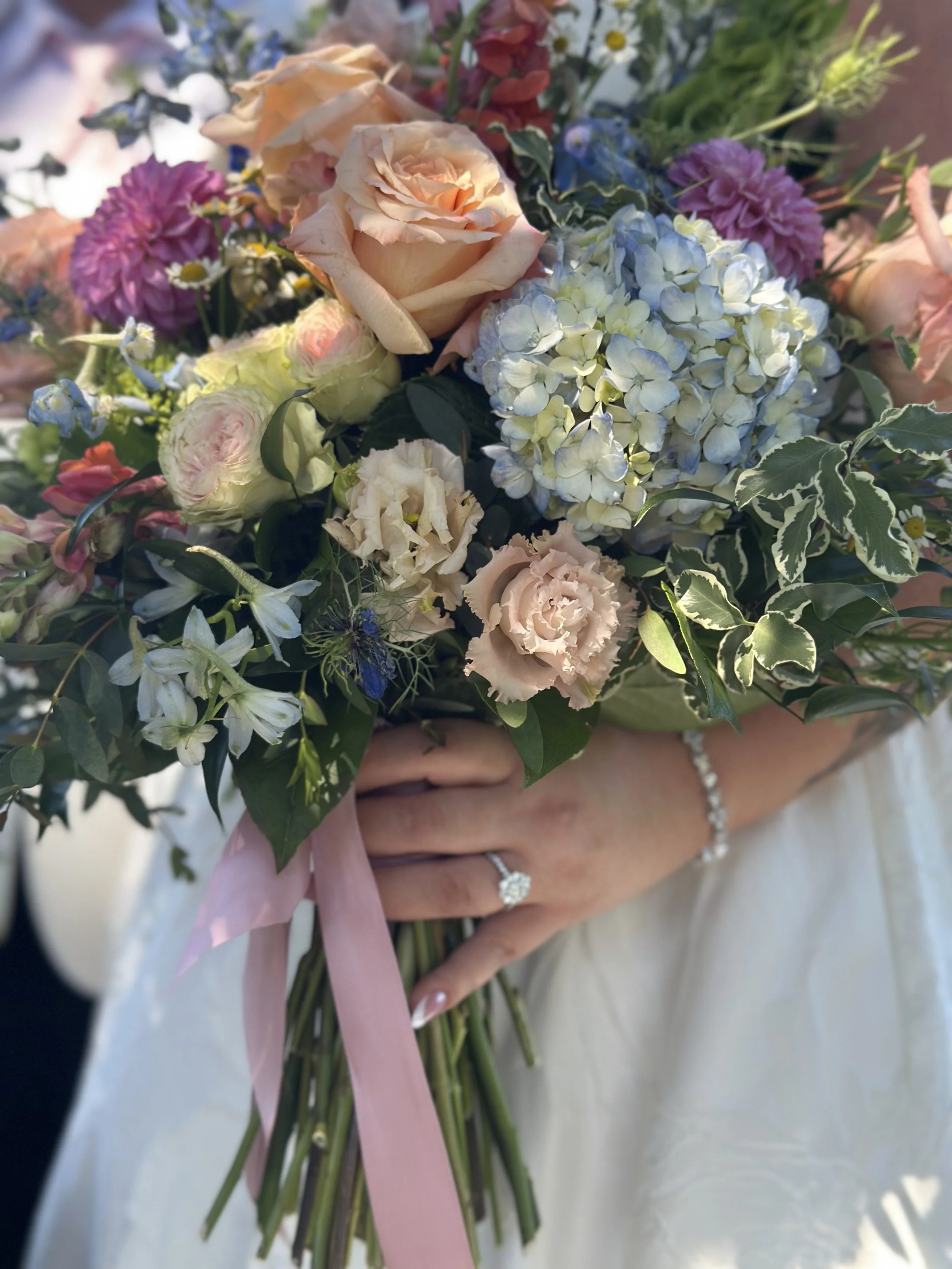 A person holding a bouquet of assorted flowers, including roses, hydrangeas, and daisies, with pink ribbon. The person is wearing a ring and bracelet.