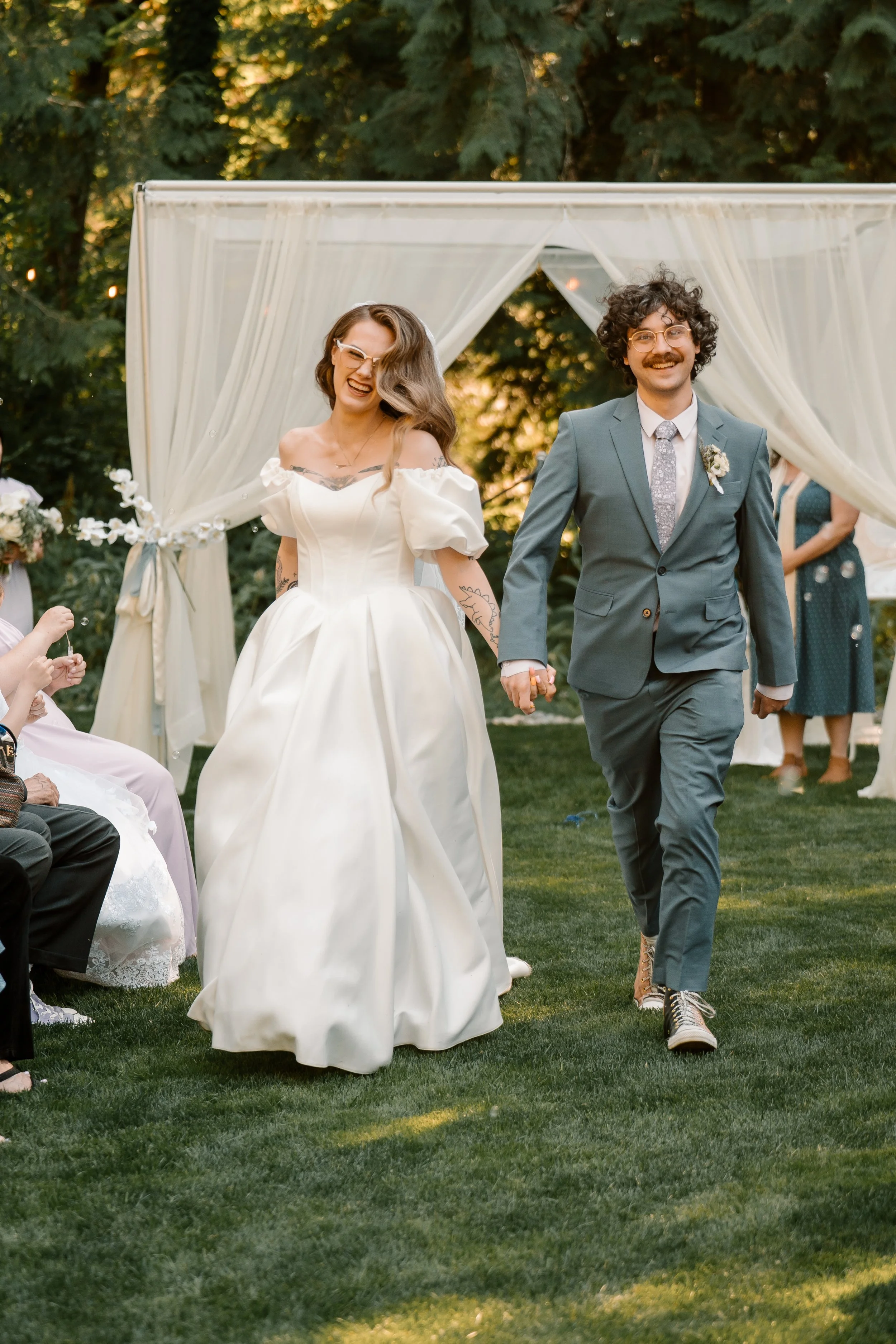 A couple, dressed in wedding attire, holding hands and walking together outdoors under a white canopy with flowing curtains, surrounded by lush greenery, during what appears to be a wedding ceremony.
