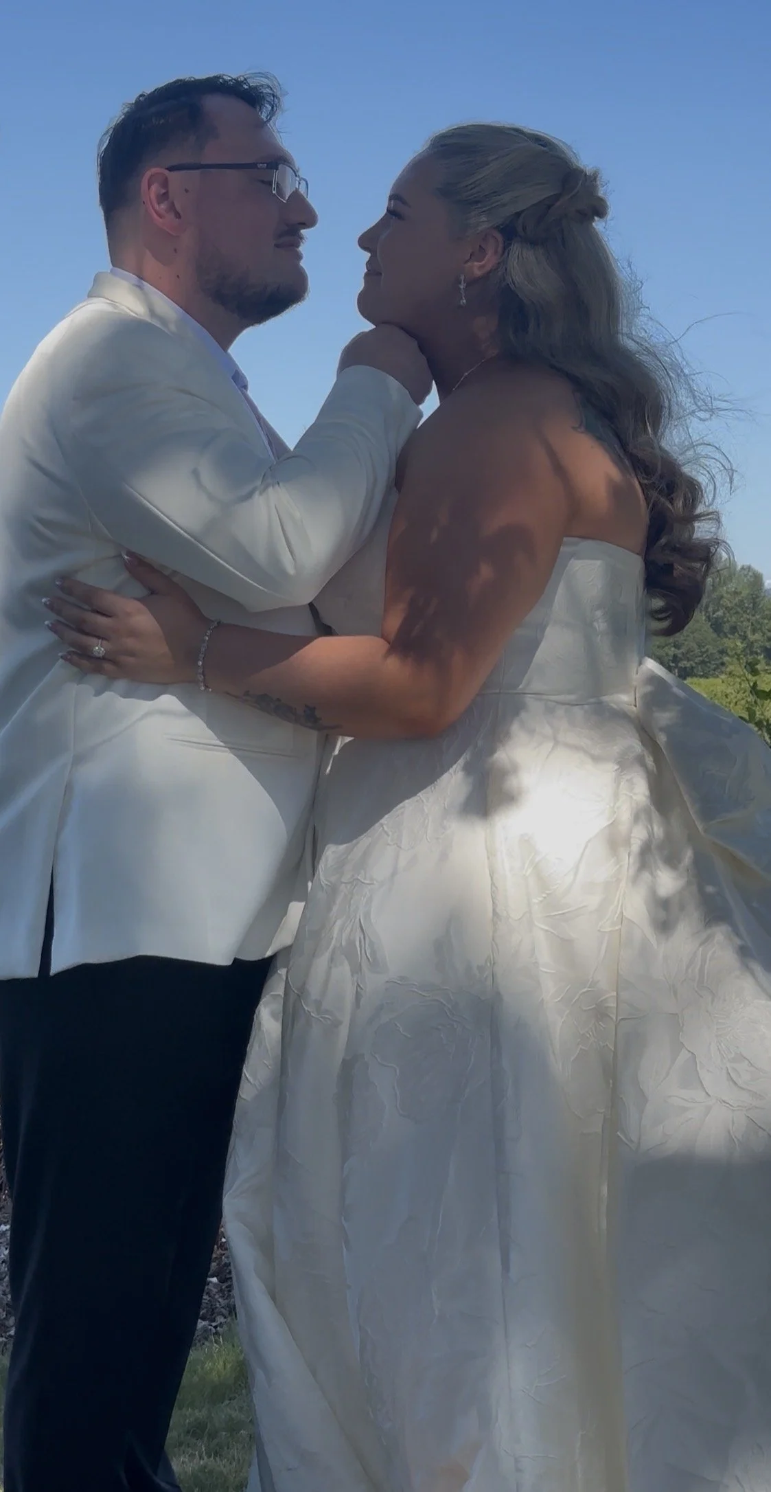 A couple dressed in wedding attire embrace outdoors under a clear blue sky, with the man gently holding the woman's face as they gaze into each other's eyes.