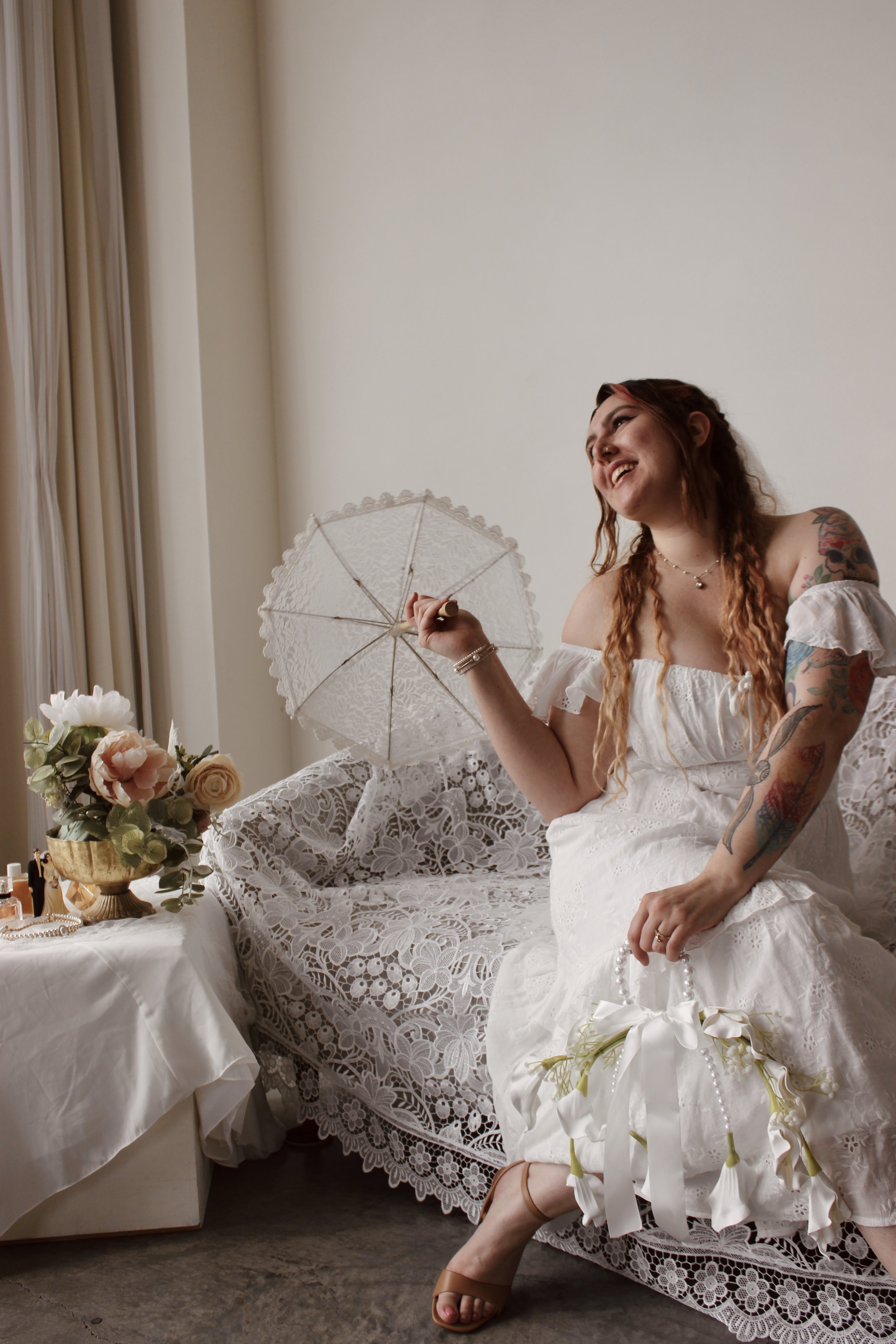 A woman in a white wedding dress with colorful tattoos on her arms, sitting on a lace-covered chair, smiling, holding a string of pearls, surrounded by flowers and wedding-related items, in a room with beige walls and curtains.