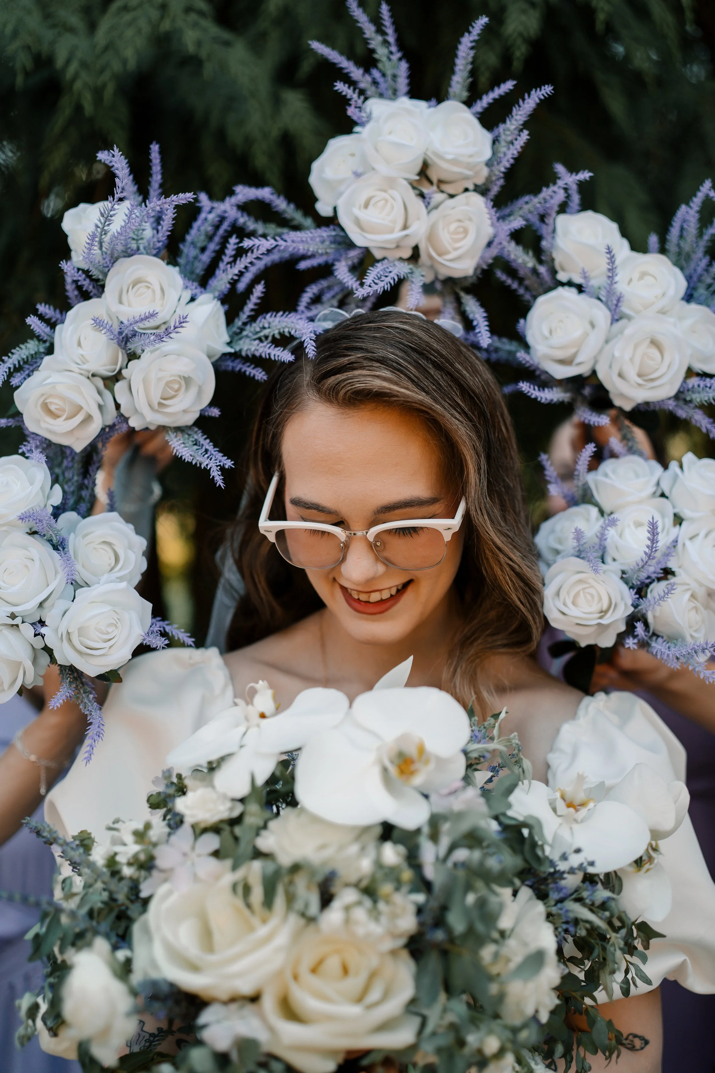 A woman wearing glasses is smiling and holding a bouquet of white flowers, with a floral crown of white roses and purple lavender around her head.
