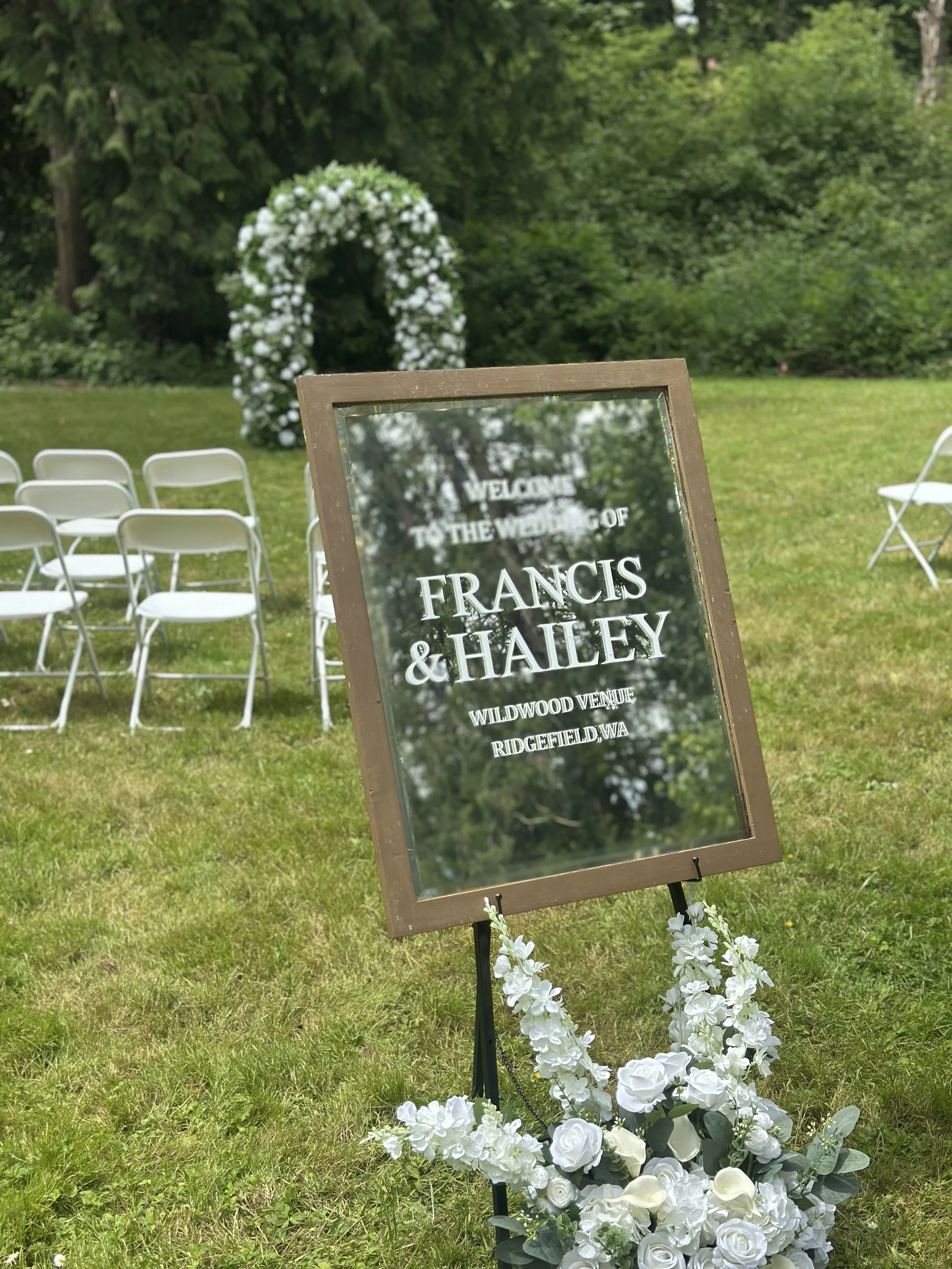 A wedding ceremony setup in a lush green outdoor setting with white chairs arranged on grass and a floral arch in the background. A sign with the names "Francis & Haley" and wedding details is in the foreground, adorned with white flowers at its base.